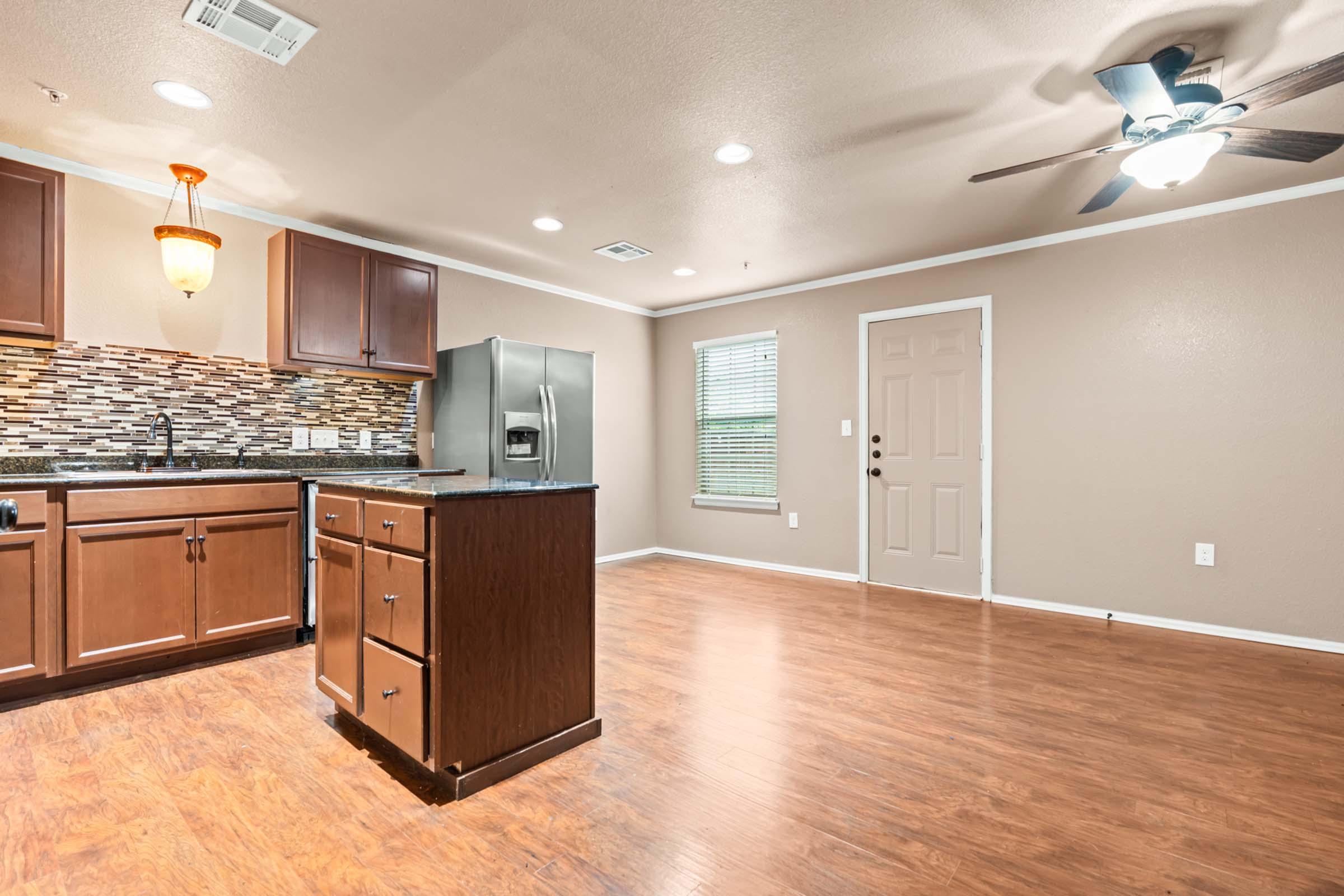 Interior view of a modern kitchen and living area. The kitchen features dark wood cabinets, a decorative backsplash, and stainless steel appliances. The living area has hardwood floors, a ceiling fan, and a door leading outside. Natural light enters through a window, enhancing the open layout of the space.