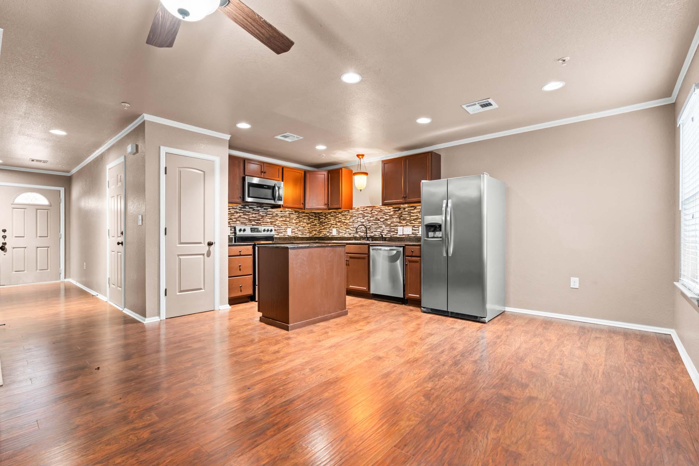 Modern kitchen and living area with wooden flooring, featuring dark wood cabinets, stainless steel appliances, a tiled backsplash, and an open layout. Brightly lit with ceiling lights, the space includes an entrance door visible on the left.