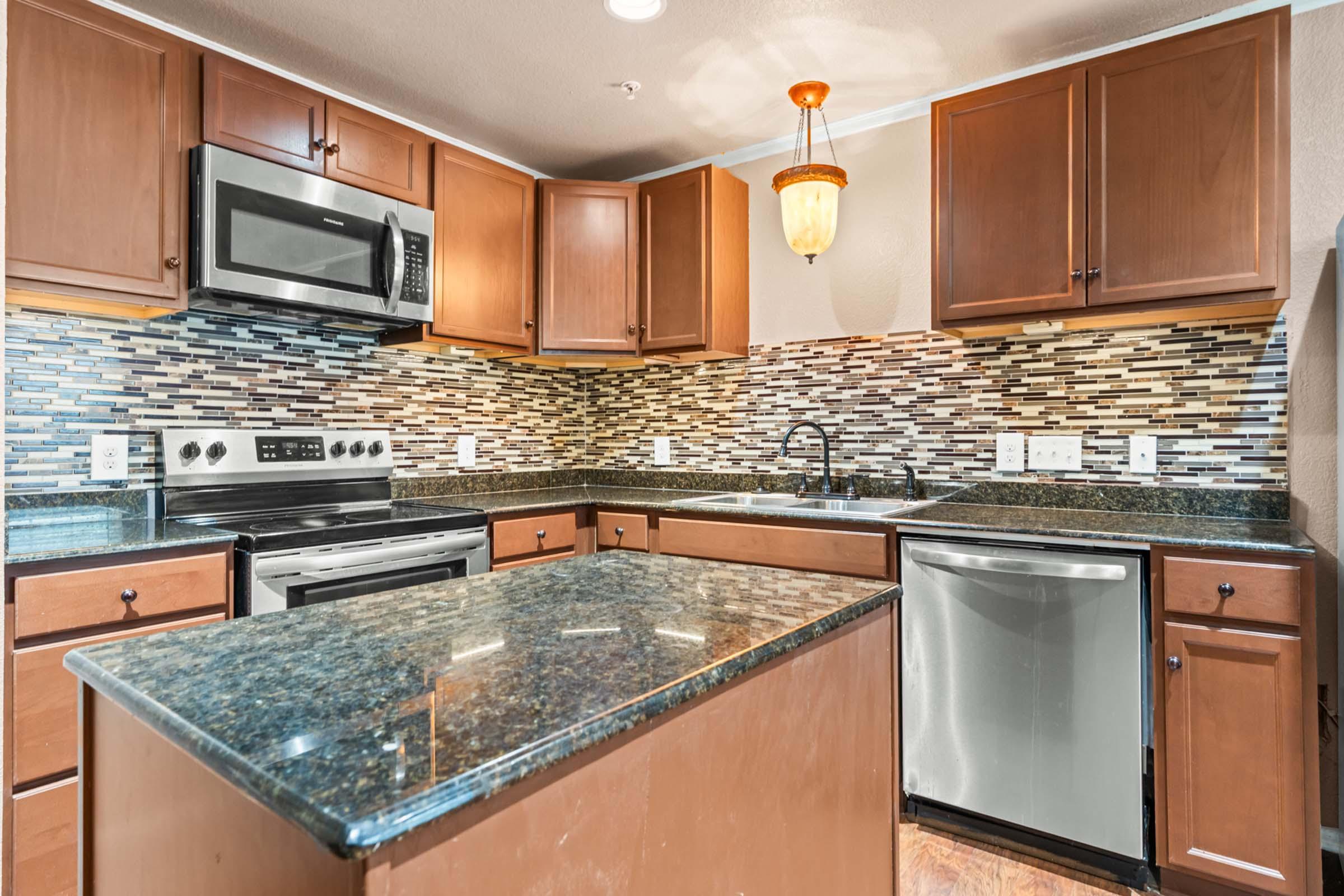 Modern kitchen featuring wooden cabinets, stainless steel appliances including a microwave and oven, a granite countertop island, and a decorative tile backsplash. The space is well-lit with an overhead pendant light.