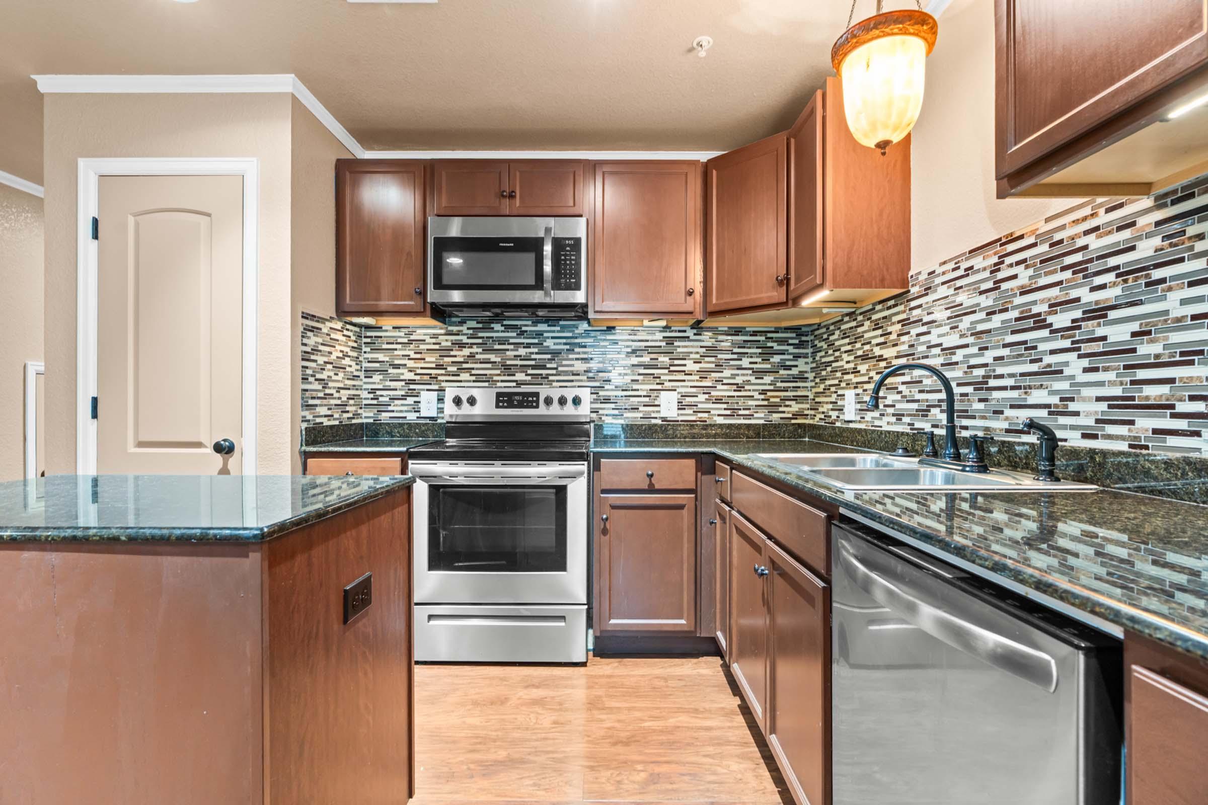 Modern kitchen featuring dark wooden cabinets, a stainless steel refrigerator, oven, and microwave. The countertops are dark granite, and a stylish backsplash of mosaic tiles adds visual interest. Includes a double sink and dishwasher. soft lighting creates a warm atmosphere.