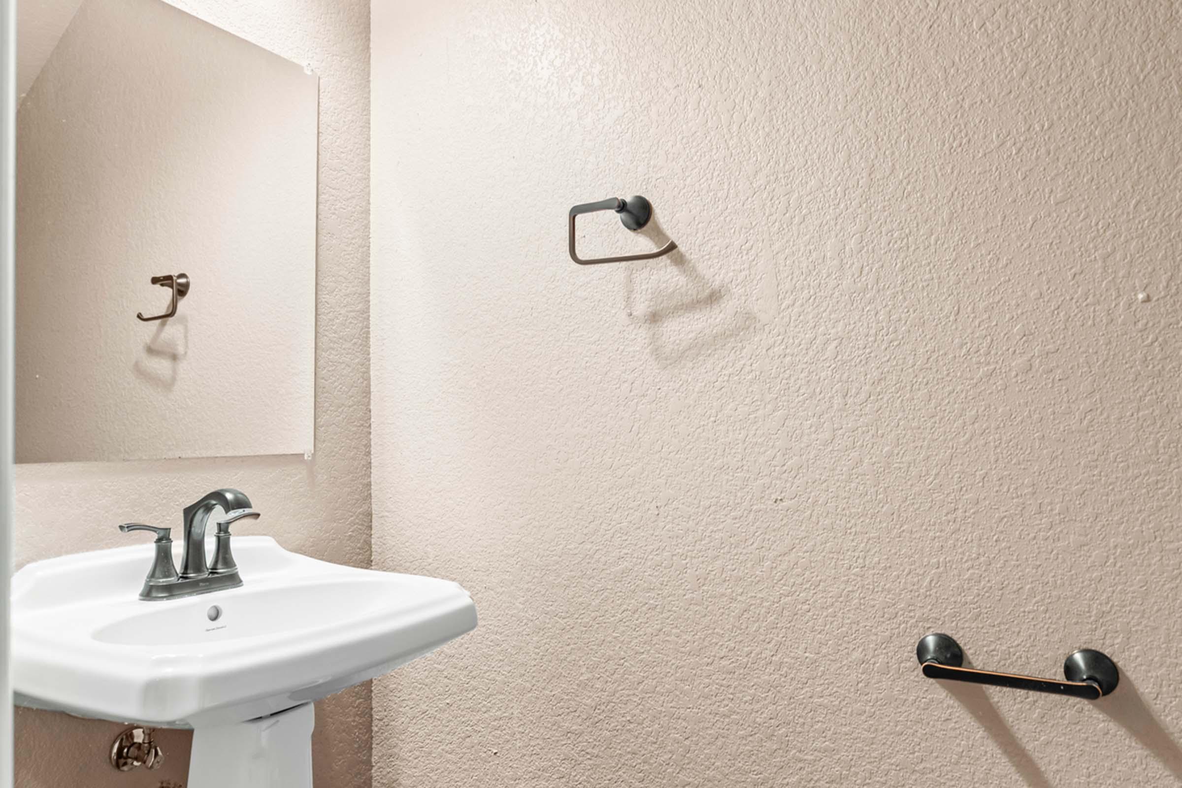 A small bathroom featuring a white sink with a modern faucet, a mirror above the sink, and beige walls. A black towel rack is mounted on the wall next to the sink, and a small towel holder is also visible. The overall design is simple and clean.