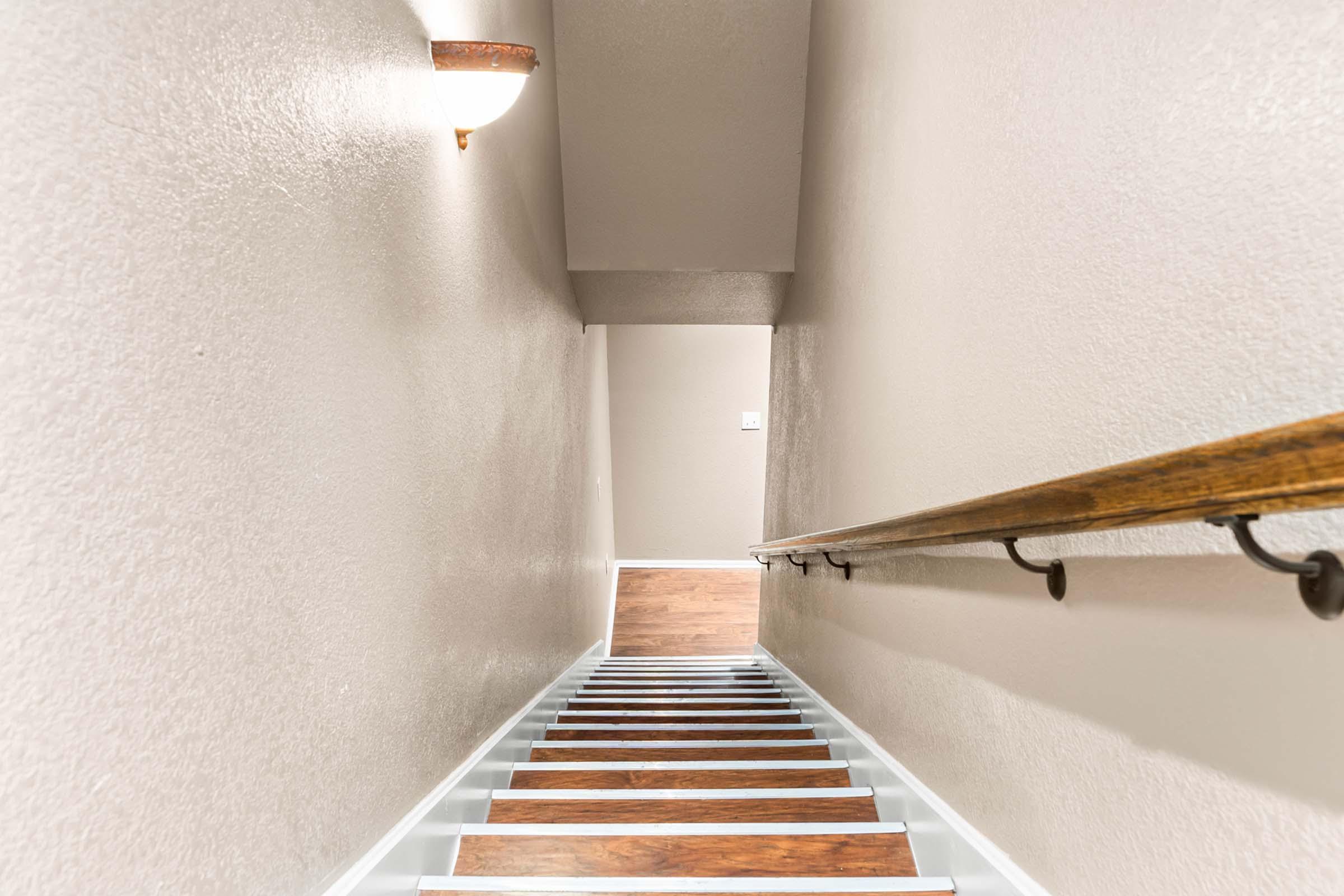 A well-lit staircase with a wooden handrail leading downward. The walls are painted in a light beige color, and the steps have a mix of wood and white finishes. A wall sconce provides soft illumination, creating a warm atmosphere in the space.