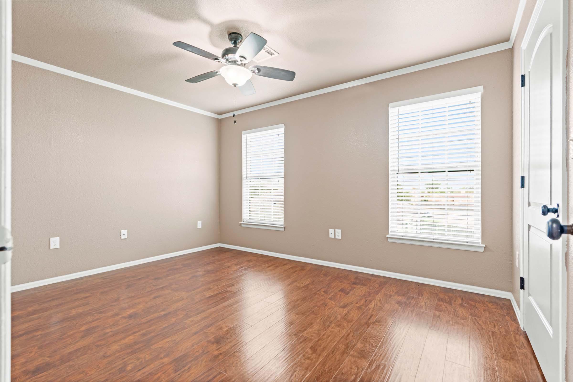 Empty room with light brown walls and wood-style flooring. Features a ceiling fan and two windows with white blinds allowing natural light. The room has one door and is free of furniture, creating a spacious and inviting atmosphere.