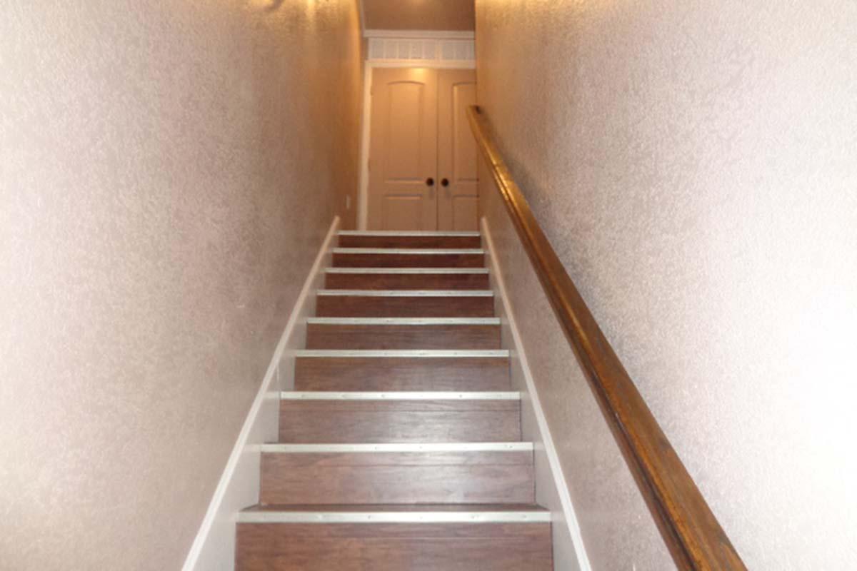 Stairs leading up a narrow hallway with wooden handrail and light-colored walls. At the top, a pair of closed double doors can be seen. The staircase has wooden steps with a light-colored trim, and the lighting creates a warm ambiance.