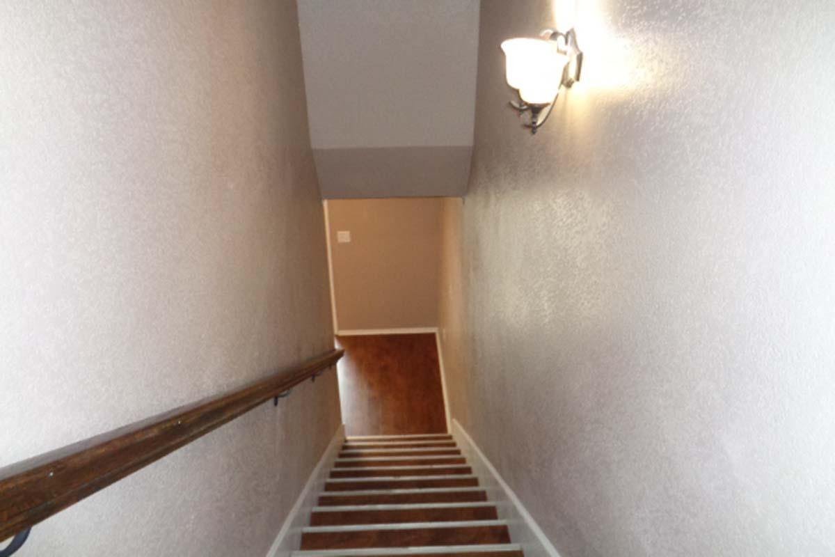 Staircase leading down with wooden railing and wall-mounted light fixture. The walls are painted in a soft color, and the flooring at the bottom is visible.