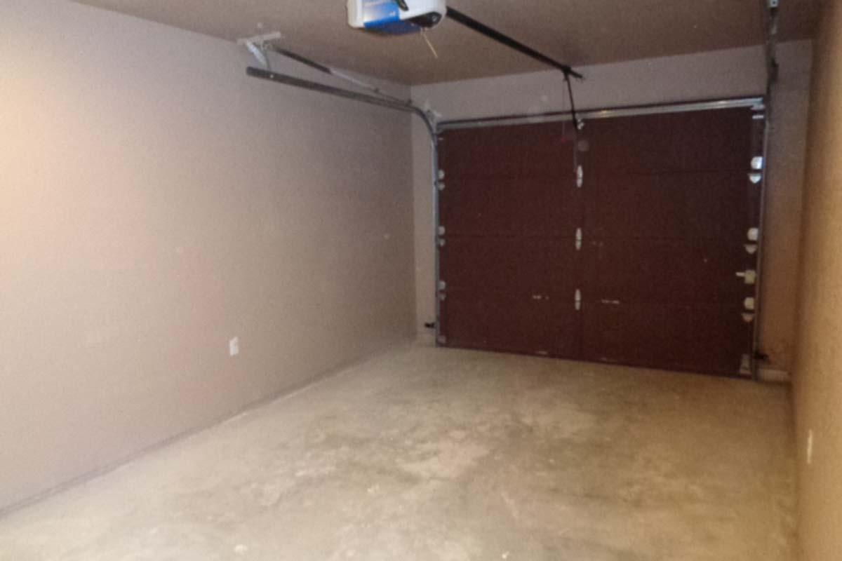 An empty garage with concrete flooring and a brown garage door. The walls are painted a light color, and a ceiling-mounted garage door opener is visible. There are no vehicles or objects present in the space.