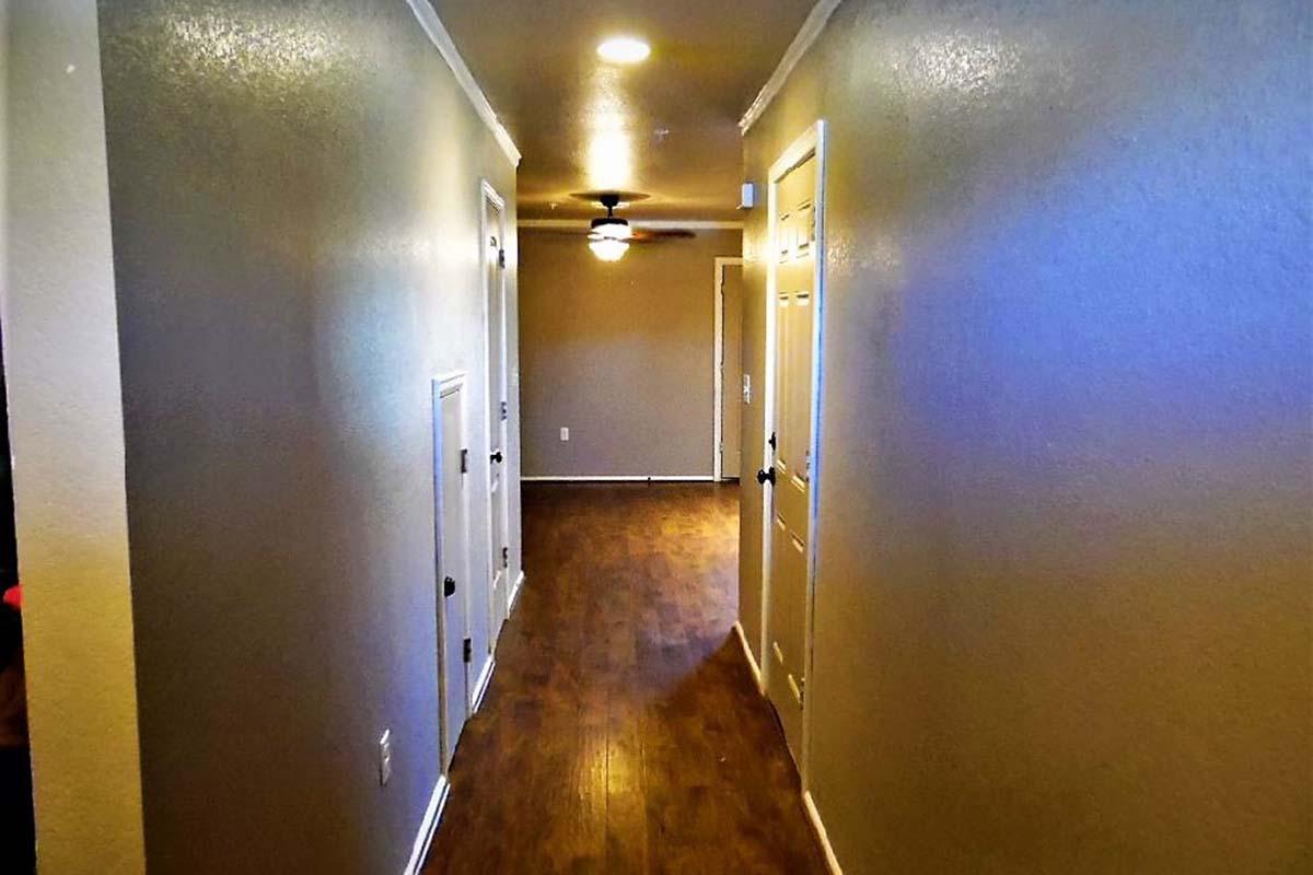 A well-lit hallway with beige walls and a wooden floor. The corridor features white doors on either side, leading to various rooms. A ceiling fan is visible at the far end, and there are small textured details on the walls, adding depth to the space. The overall atmosphere is warm and inviting.