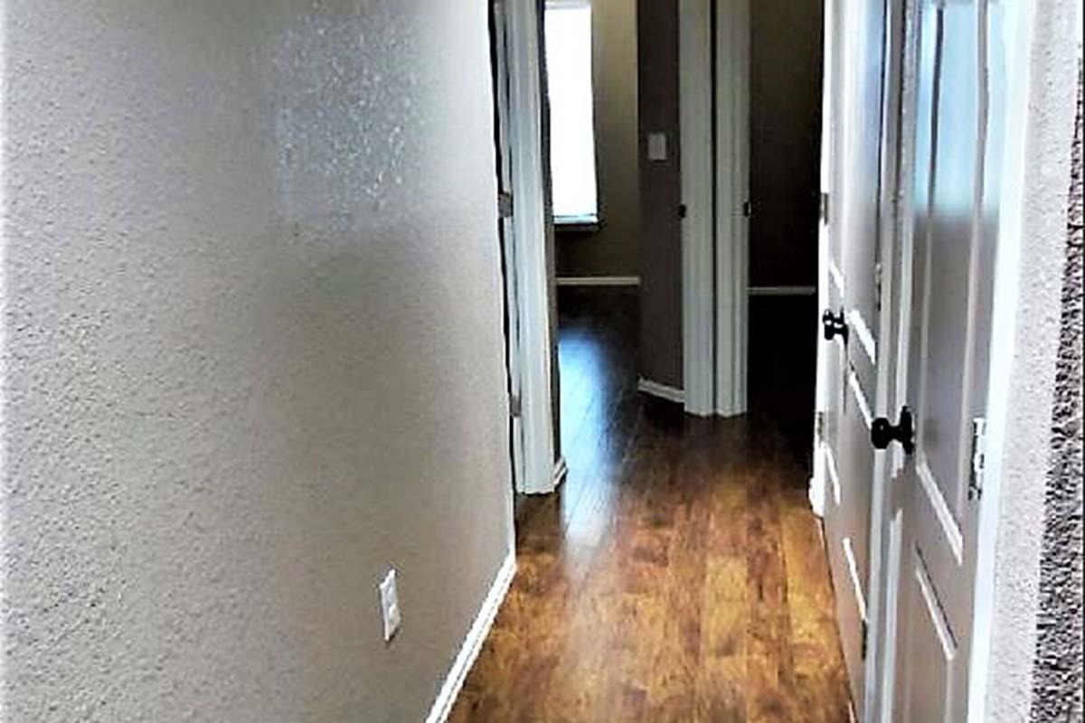 A narrow hallway featuring light brown walls and wooden flooring. There are two doors on the left, with closed white doors, and a window in the distance allowing natural light to brighten the space. The hallway leads to additional rooms beyond the visible area.