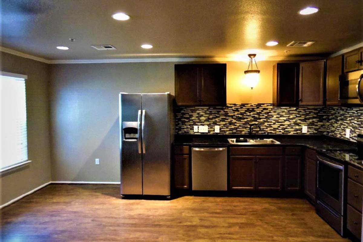 A modern kitchen featuring dark wood cabinets, stainless steel appliances including a refrigerator and dishwasher, and a black countertop. The backsplash is made of mosaic tiles in shades of gray and black. The room has recessed lighting and a window allowing natural light to enter.