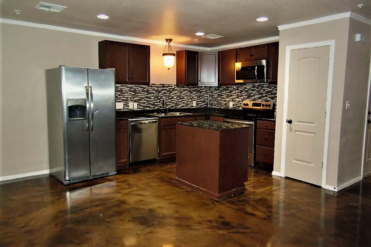 Modern kitchen featuring dark wood cabinets, stainless steel appliances including a refrigerator, dishwasher, and oven. The countertops are dark, with a decorative backsplash. The floor has a polished finish. There is ample lighting from the ceiling fixture and natural light from the doorway.