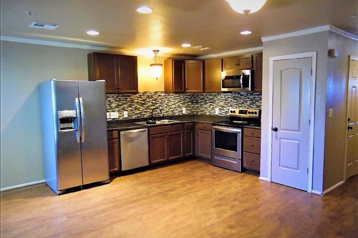 Modern kitchen featuring stainless steel appliances, including a refrigerator, oven, and dishwasher. The cabinets are dark wood, and the backsplash includes a mix of tiles. The floor has a wood-like finish, and there's a light fixture on the ceiling, creating a warm ambiance.