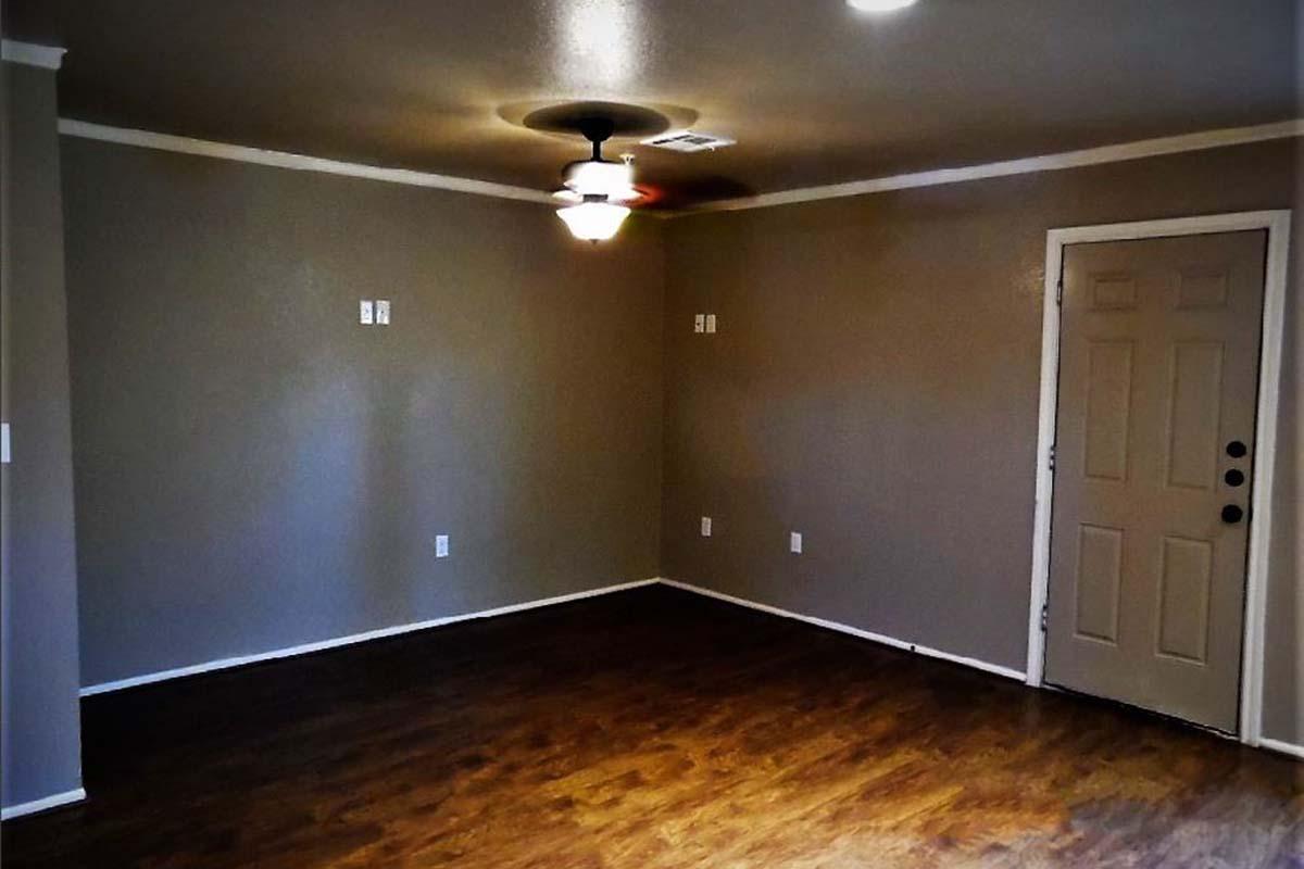 A spacious, empty room featuring light brown walls and dark wooden flooring. There is a ceiling fan in the center, and the room has a single door on one side. The walls are mostly bare, with a few electrical outlets visible. Soft lighting adds warmth to the space.