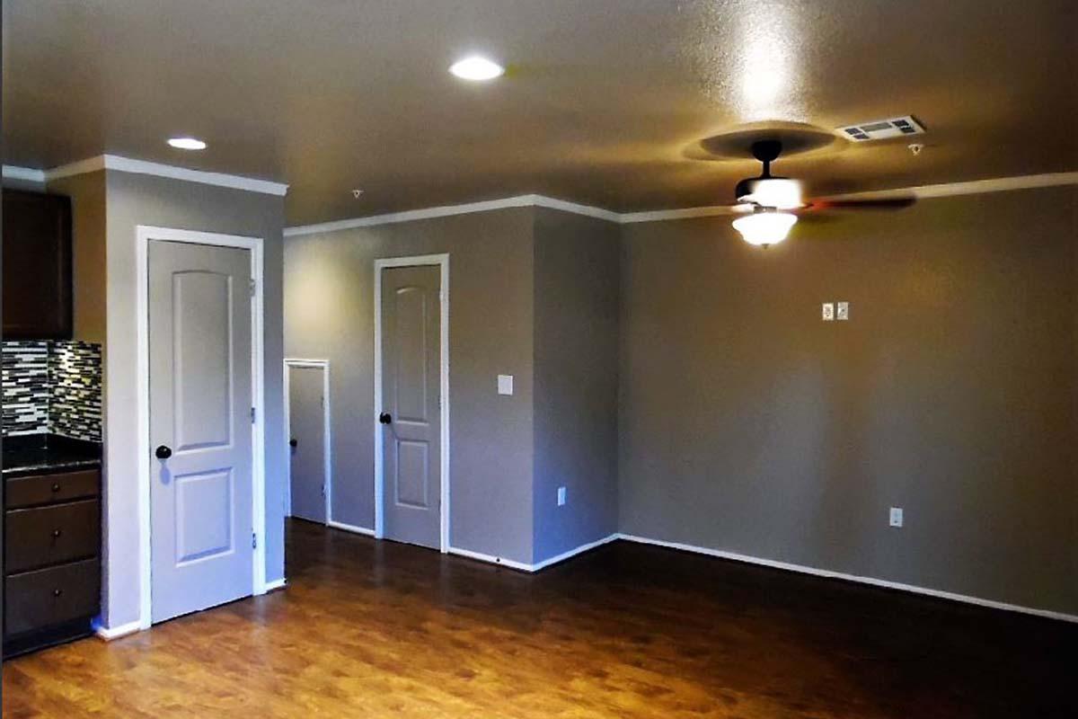 A spacious room featuring light-colored walls, a ceiling fan, and wood laminate flooring. There are two doors leading to other areas, and a kitchen area with dark cabinets and a decorative backsplash on one side. The room is well-lit with recessed ceiling lights.