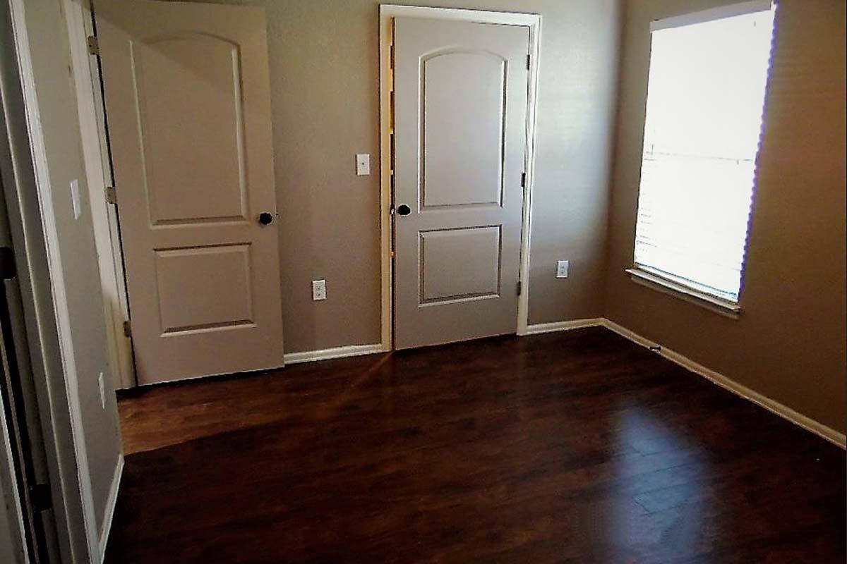 A sparsely furnished room with light brown walls and laminate wood flooring. It features two closed doors on the left side, a window with blinds on the right, and no furniture or decorations in the space, creating a minimalist appearance.