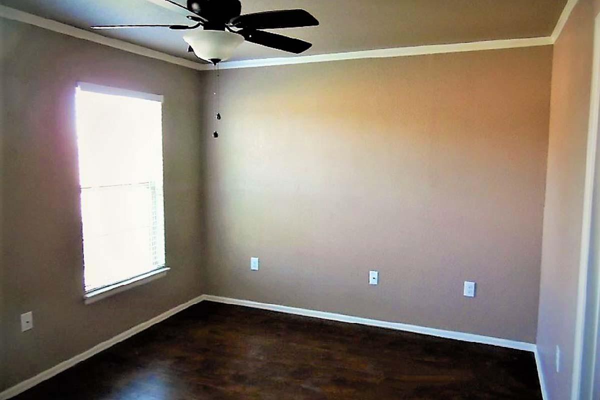 Empty room with beige walls, a ceiling fan, and a window letting in natural light. The floor is wooden, and there are electrical outlets on the walls. The space appears clean and ready for furniture.