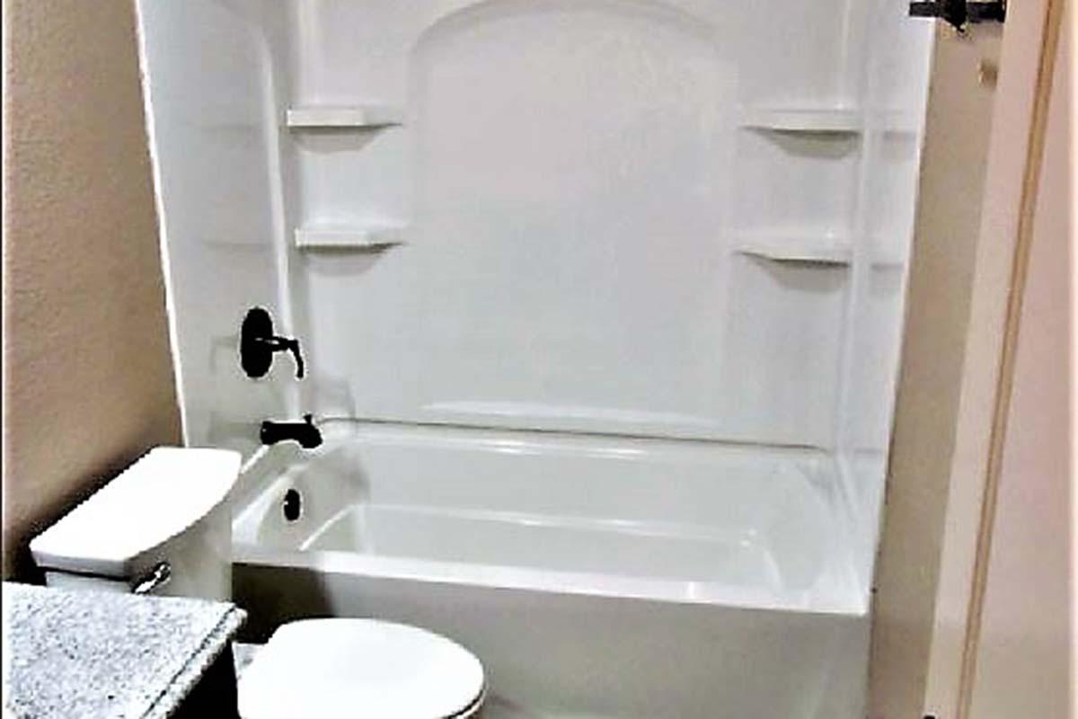 A clean, modern bathroom featuring a white bathtub with a shower attachment, a toilet, and a small sink with a granite countertop. The walls are painted beige, and there are built-in shelves in the bathtub enclosure for storage.