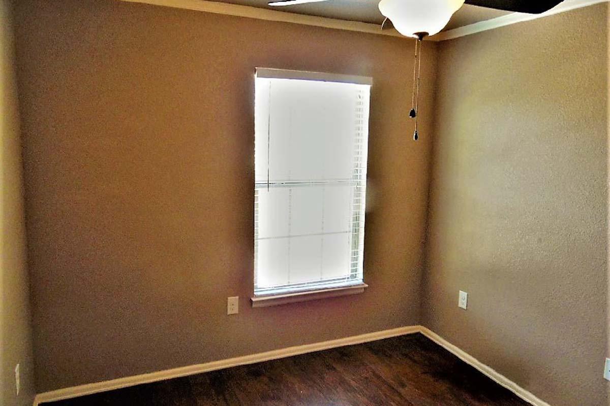 Empty room with light brown walls and a ceiling fan. A window with white blinds lets in natural light. The floor is dark wood, and there are electrical outlets on the walls. The space appears clean and ready for furnishing.