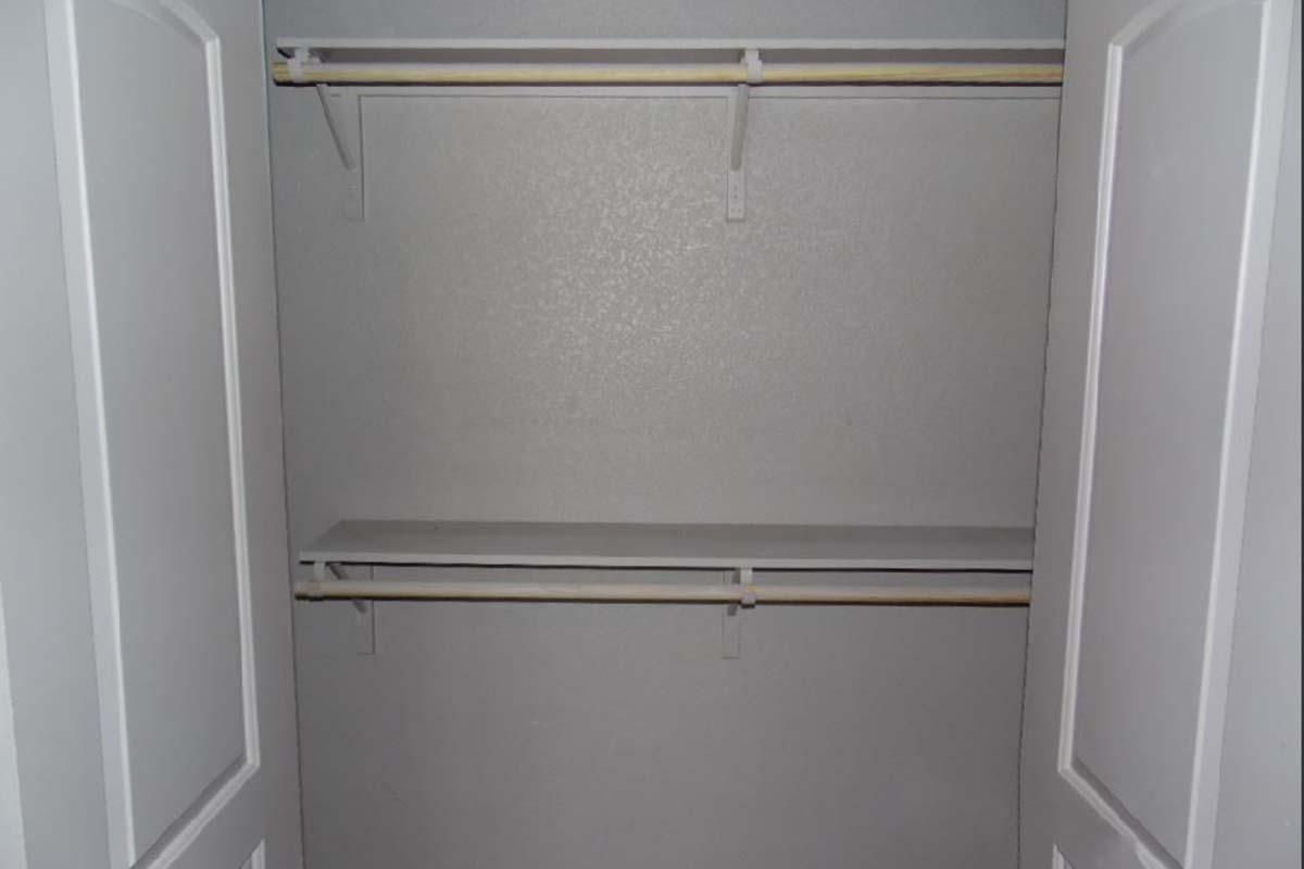 An empty closet with two horizontal shelves on the upper and lower parts, featuring a plain, light-colored wall. The closet doors are open, revealing the lack of any stored items or clothing.