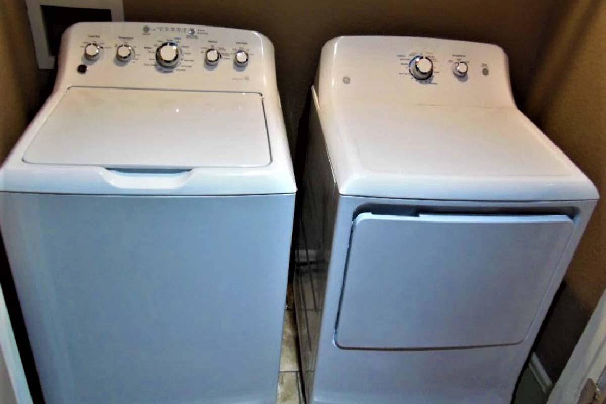 A white washing machine and a white dryer placed side by side in a laundry room. The washing machine features multiple cycle settings and a large lid, while the dryer has a control panel with knobs for temperature and drying options. The walls are a neutral color, and the floor has tiled patterns.