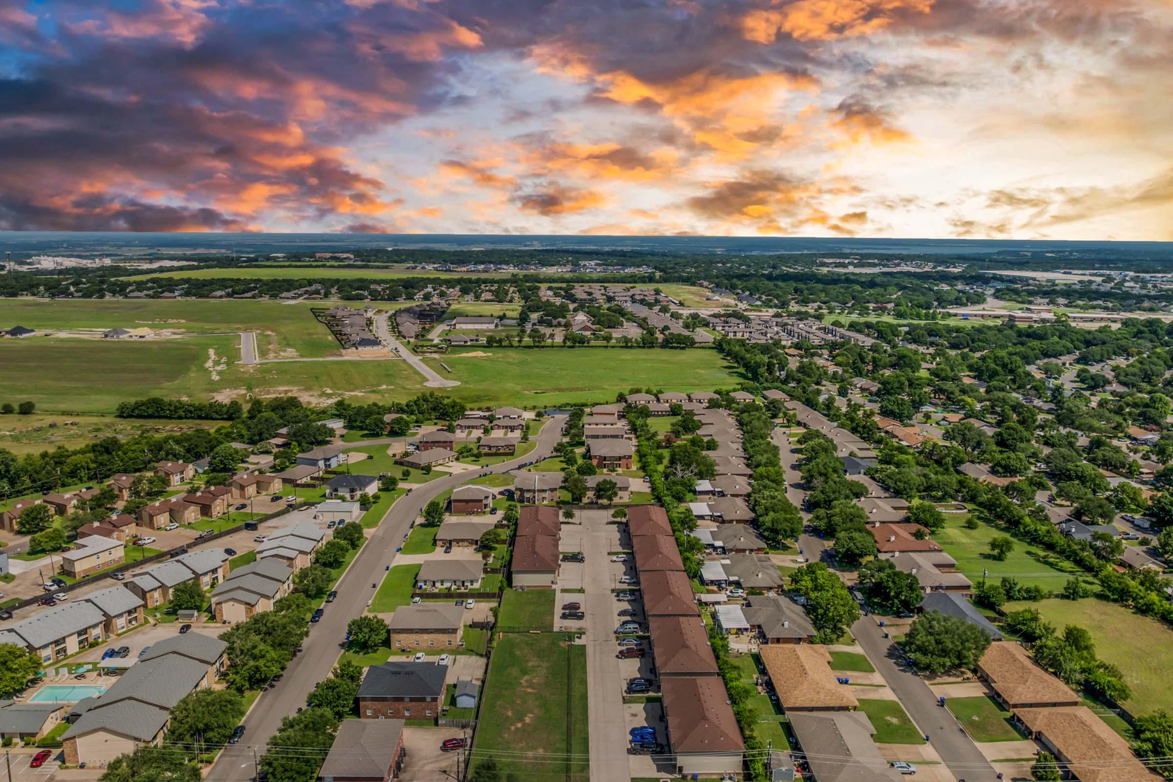 An aerial view of a suburban neighborhood featuring rows of houses with varying roof colors, green lawns, and streets. In the background, expansive green fields and a vibrant sunset sky with orange and purple hues create a picturesque landscape.