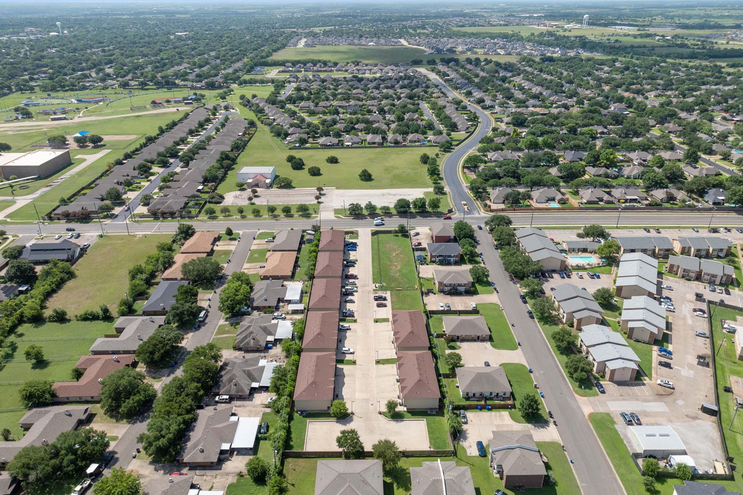 Aerial view of a suburban neighborhood featuring rows of houses, green spaces, and streets. The landscape includes well-maintained lawns, parked cars, and a mix of residential buildings. In the distance, additional homes and open areas are visible, showcasing a typical community layout.