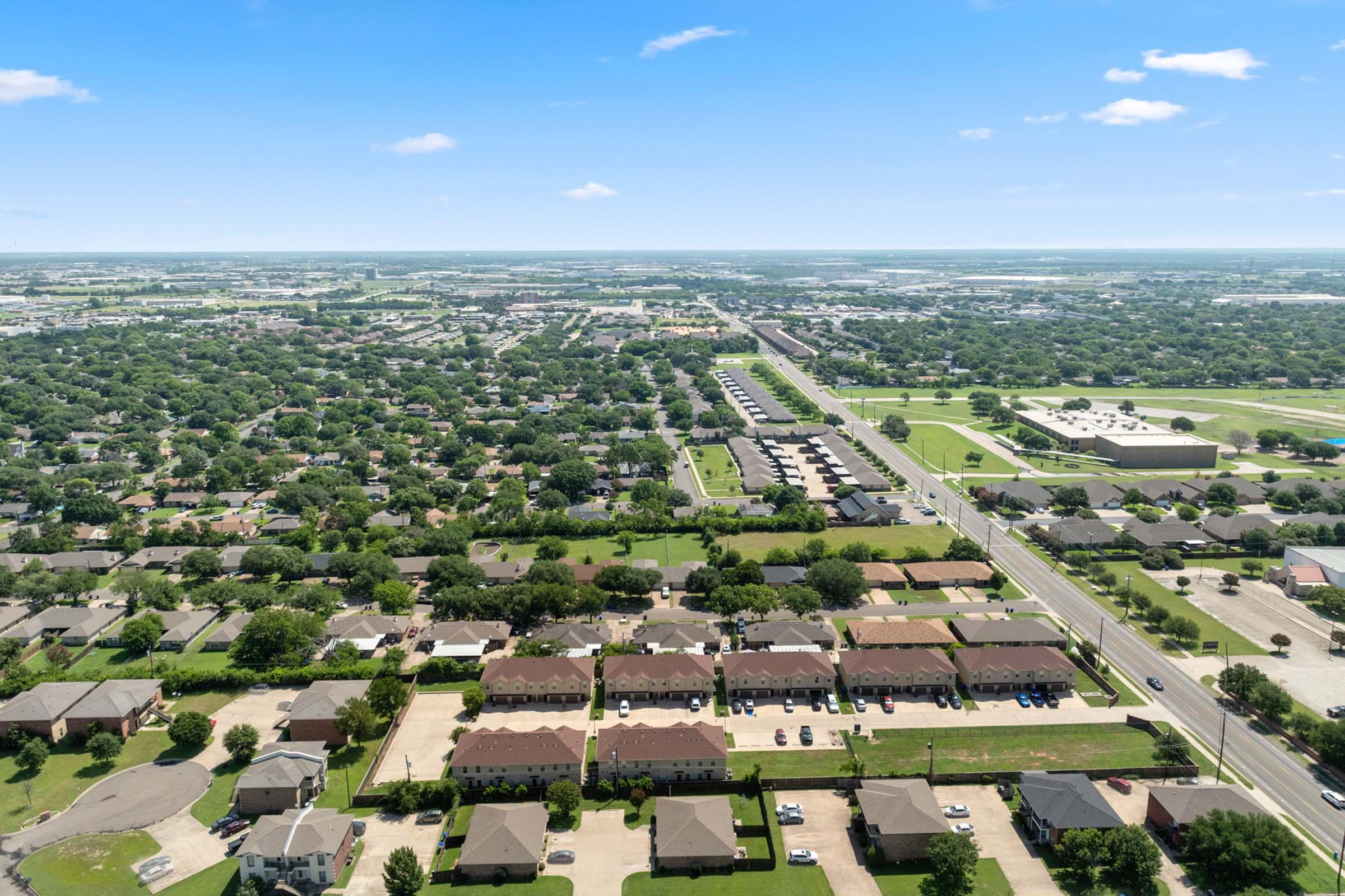 Aerial view of a suburban area featuring neatly arranged residential buildings, green lawns, and tree-lined streets. In the background, a highway and additional commercial properties are visible, surrounded by expansive fields and open spaces under a clear blue sky.