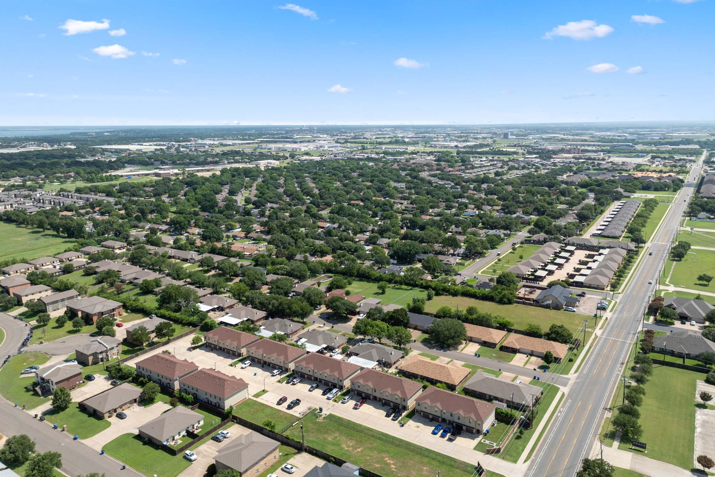 Aerial view of a suburban neighborhood with a mix of single-family homes and apartments. Lush green trees and neatly landscaped lawns are visible, along with roads and parking areas. In the distance, city buildings can be seen against a clear blue sky.