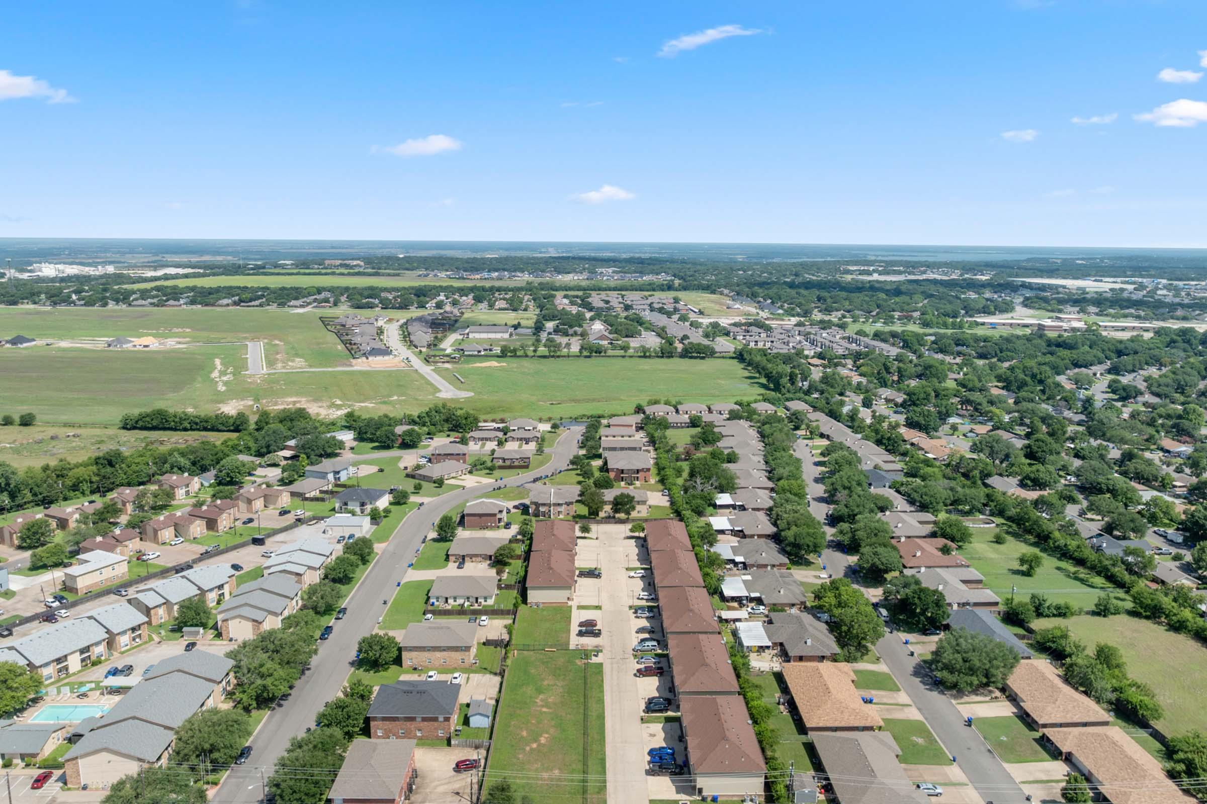 Aerial view of a suburban area featuring a network of residential streets lined with houses, green spaces, and a horizon stretching into the distance. Fields and further developments are visible, showcasing a mix of urban and rural landscapes under a clear blue sky.