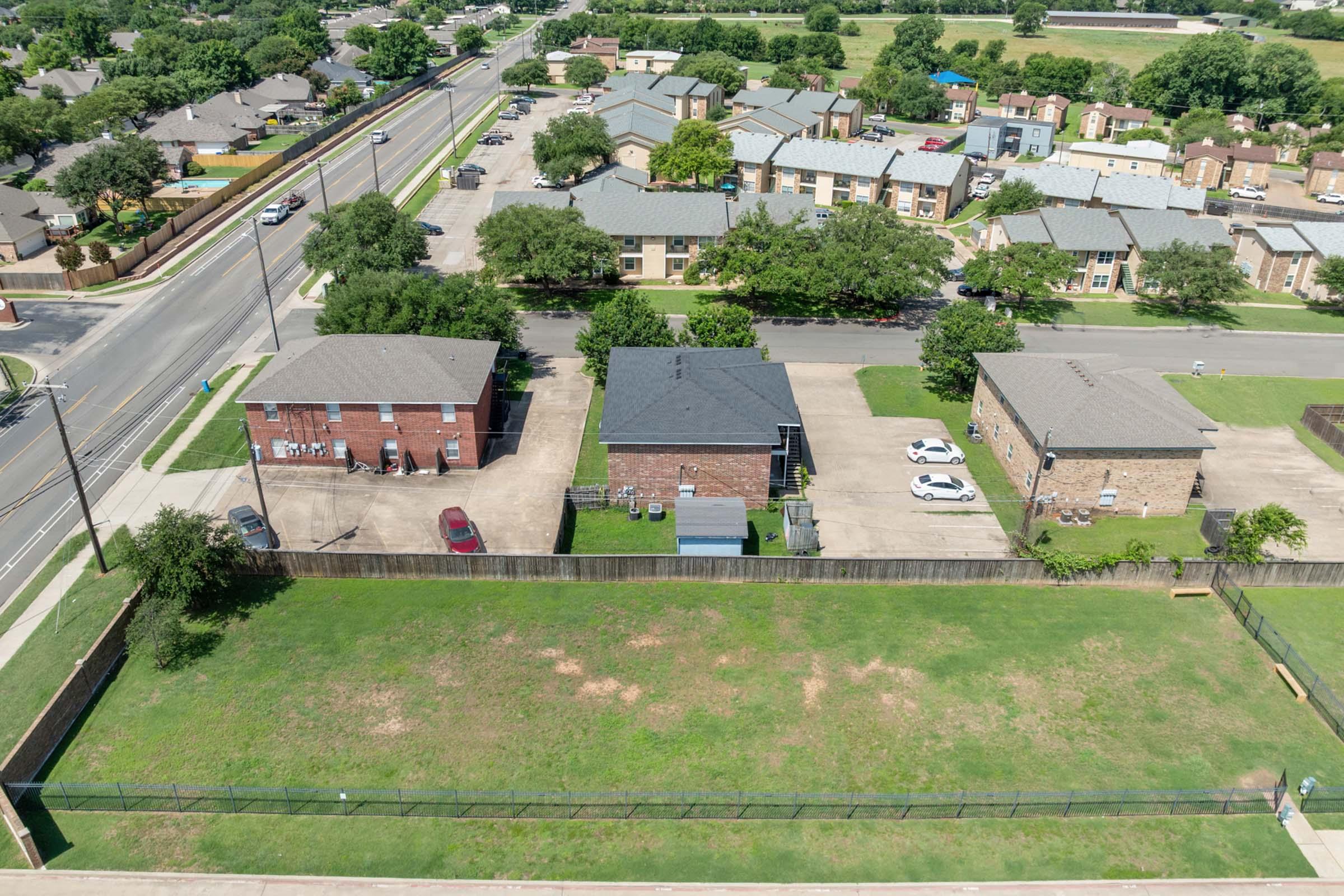 Aerial view of a residential area featuring several low-rise apartment buildings and homes. A green, grassy lot is in the foreground, while parked cars are visible in the driveways. A roadway runs alongside the neighborhood, with trees lining the streets. Nearby is a visible train track.