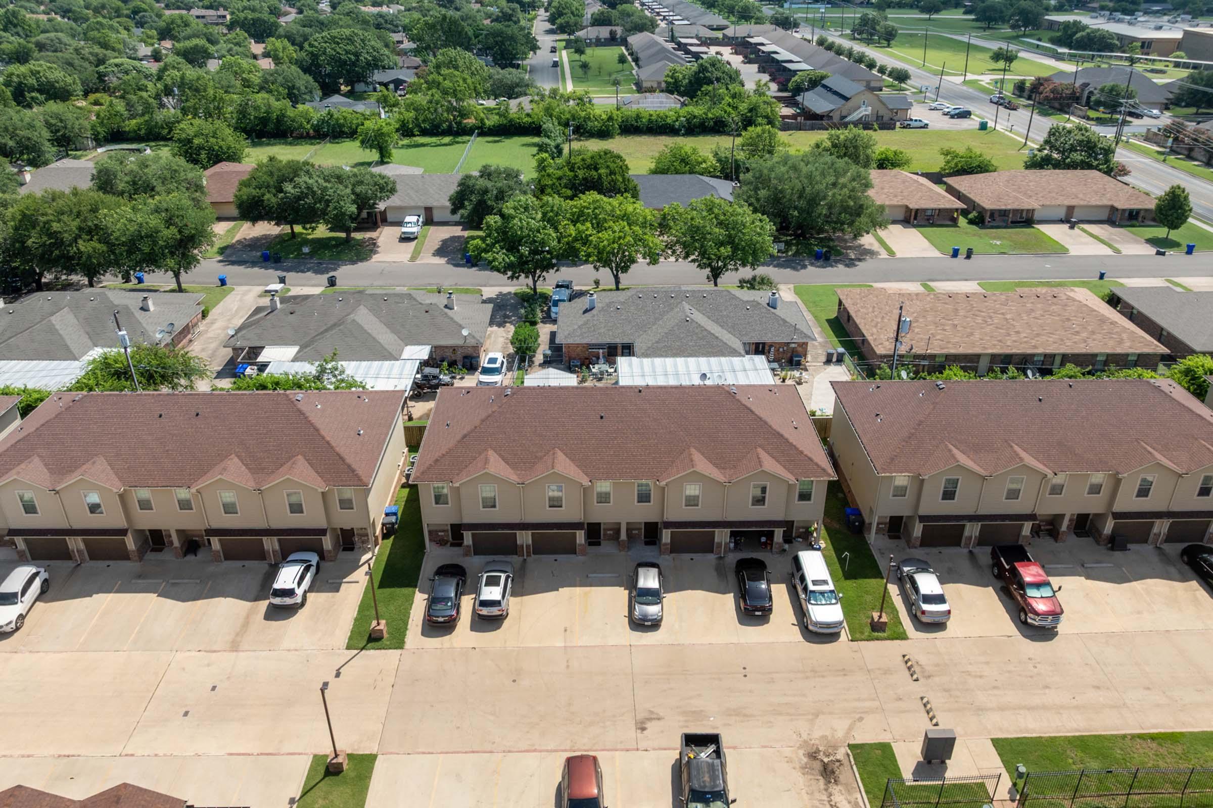 Aerial view of a residential neighborhood featuring rows of townhouses with sloped roofs, parked cars in front, and green lawns. In the background, additional homes and trees are visible, indicating a suburban setting. The layout suggests a well-maintained area with driveways and sidewalks.