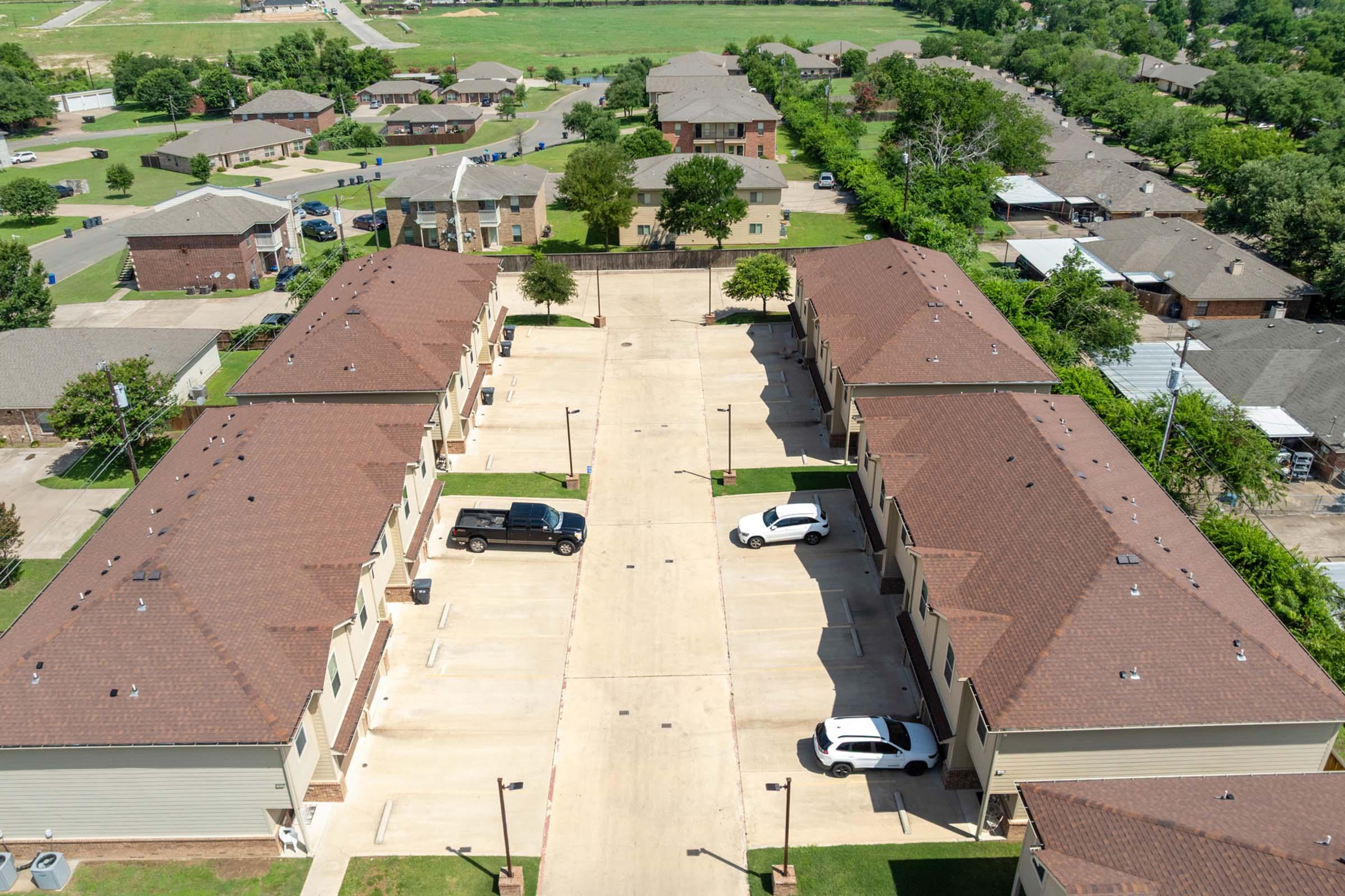 Aerial view of a residential complex featuring multiple two-story buildings arranged in a U-shape around a central parking area. There are several parked cars, green lawns, and trees lining the walkways. The surrounding area includes additional residential properties and open green spaces.