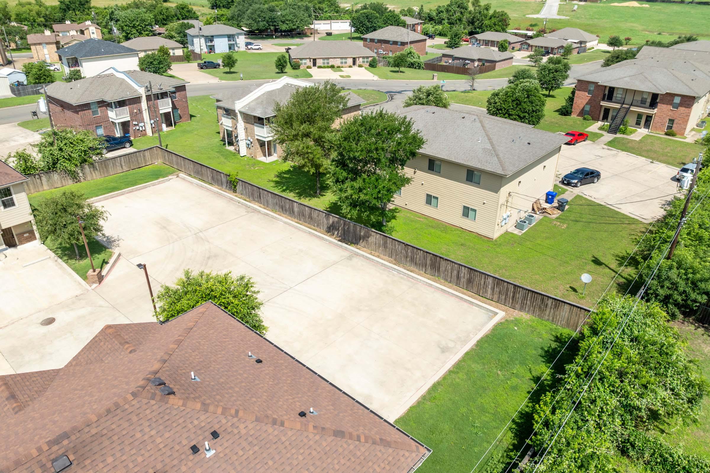 Aerial view of a residential neighborhood featuring several two-story apartment buildings, green lawns, and a fenced courtyard area. The scene includes parked cars, trees, and a paved parking lot. The layout suggests a well-maintained community with open spaces and nearby greenery.
