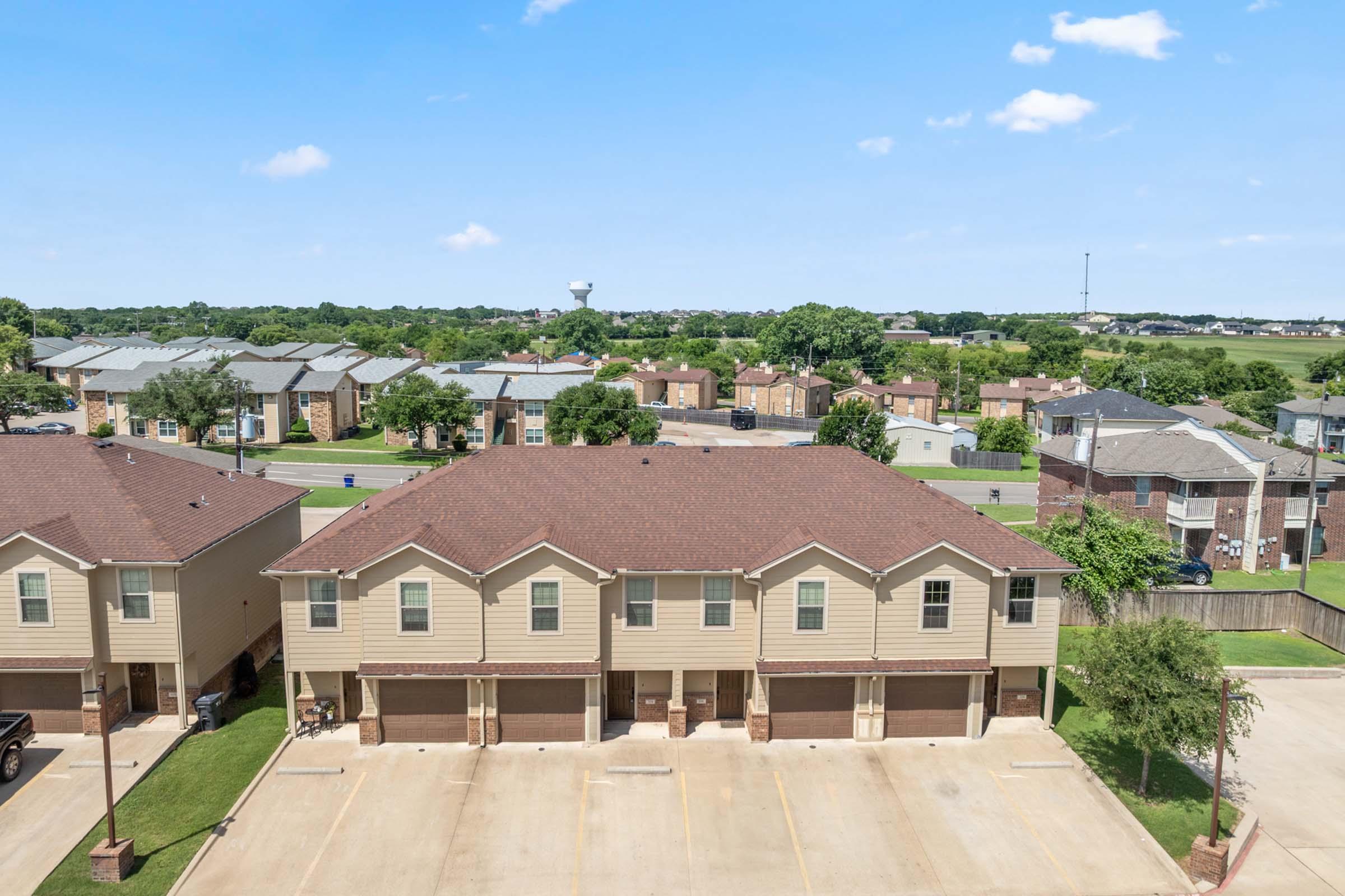 Aerial view of a residential area showcasing several two-story townhouses with brown roofs. In the background, various buildings and a water tower are visible, surrounded by greenery under a blue sky with scattered clouds. Parking spaces are in front of the townhouses.