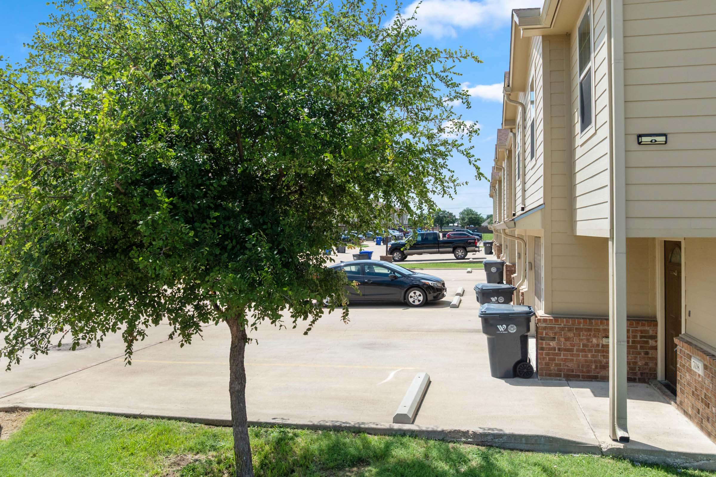 A tree is positioned in the foreground, beside an apartment complex. Visible are parked cars in a parking lot, with garbage bins near the building's entrance. The scene is set under a clear blue sky.