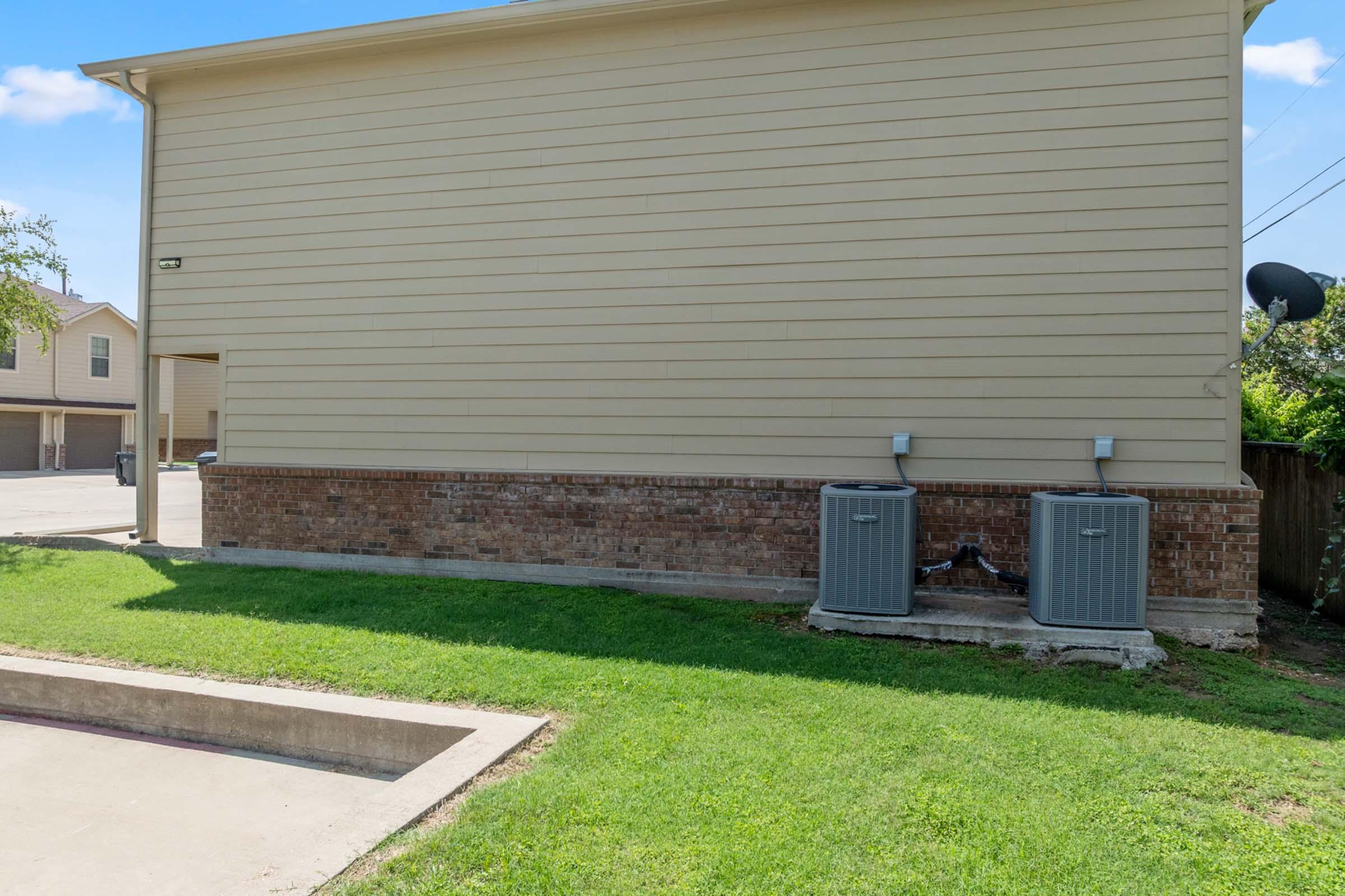 Two air conditioning units sit against the side of a beige house with a brick base. A small grassy area is in front of the units, and a concrete slab is visible nearby. The background features a clear blue sky and a few neighboring buildings.