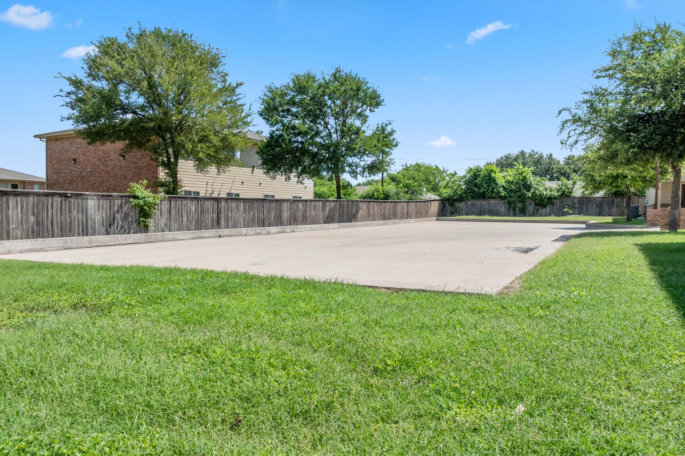 A large, vacant concrete area surrounded by green grass and trees, with a wooden fence in the background. The sky is clear with a few clouds, and there are residential buildings visible on the left side of the image.
