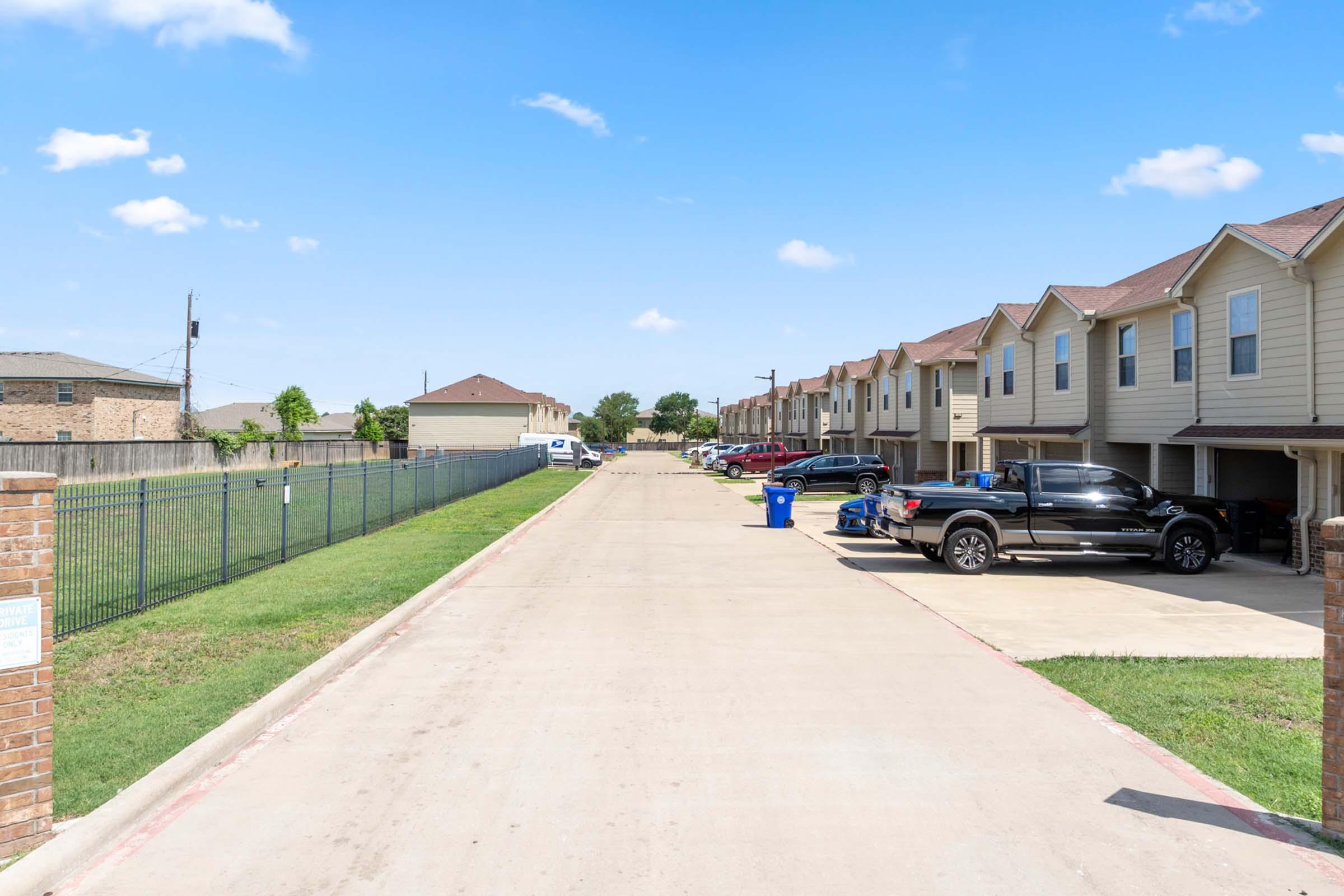 A wide view of a residential street lined with townhouses. The pavement is clear, with several parked cars, including a black pickup truck. In the background, there are more houses and a fenced area. The sky is bright blue with a few fluffy clouds, creating a sunny day atmosphere.