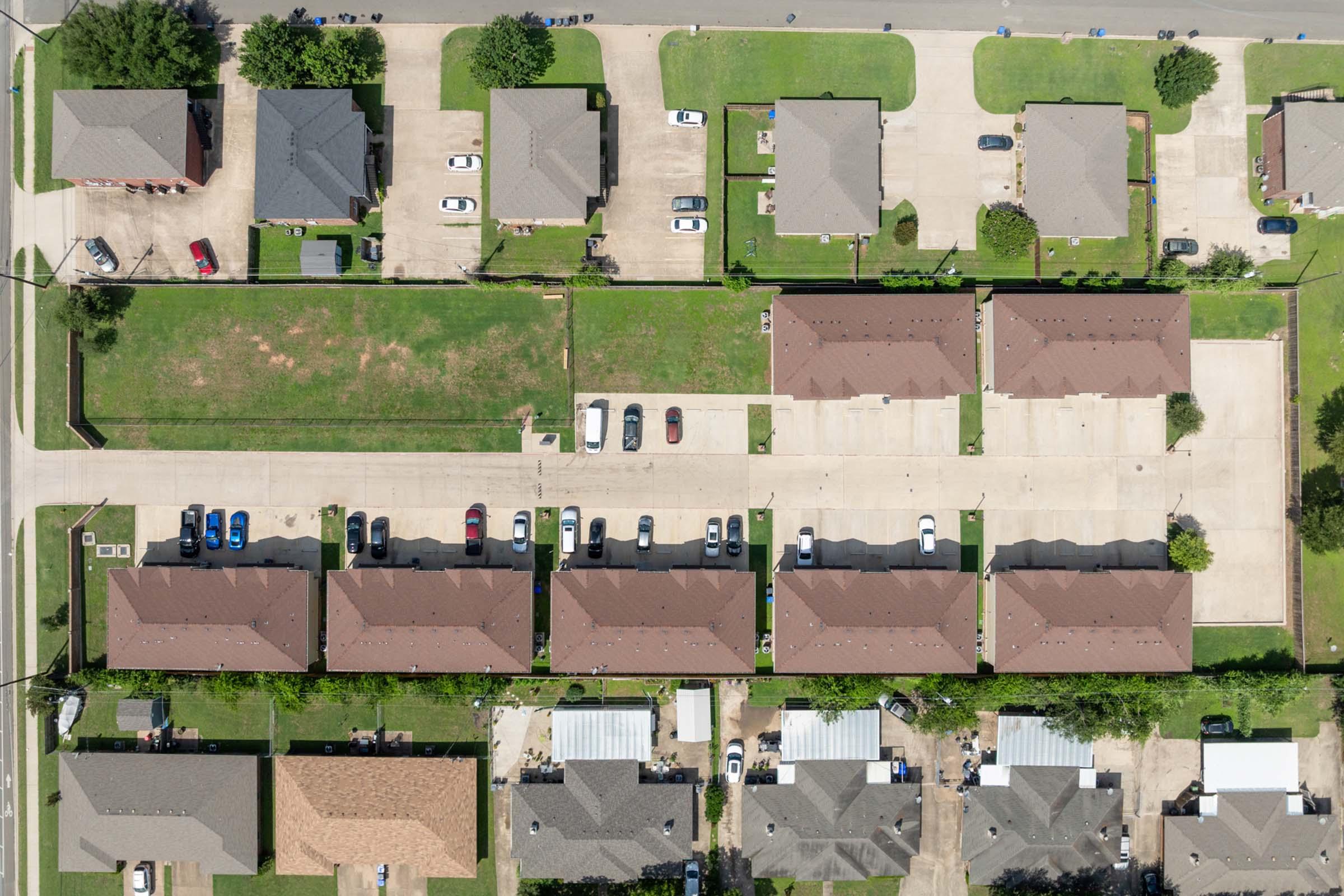 Aerial view of a residential area showcasing several single-family homes with pitched roofs, green lawns, and a parking area. The layout includes multiple rows of houses, some with parked cars, surrounded by well-maintained yards and streets.