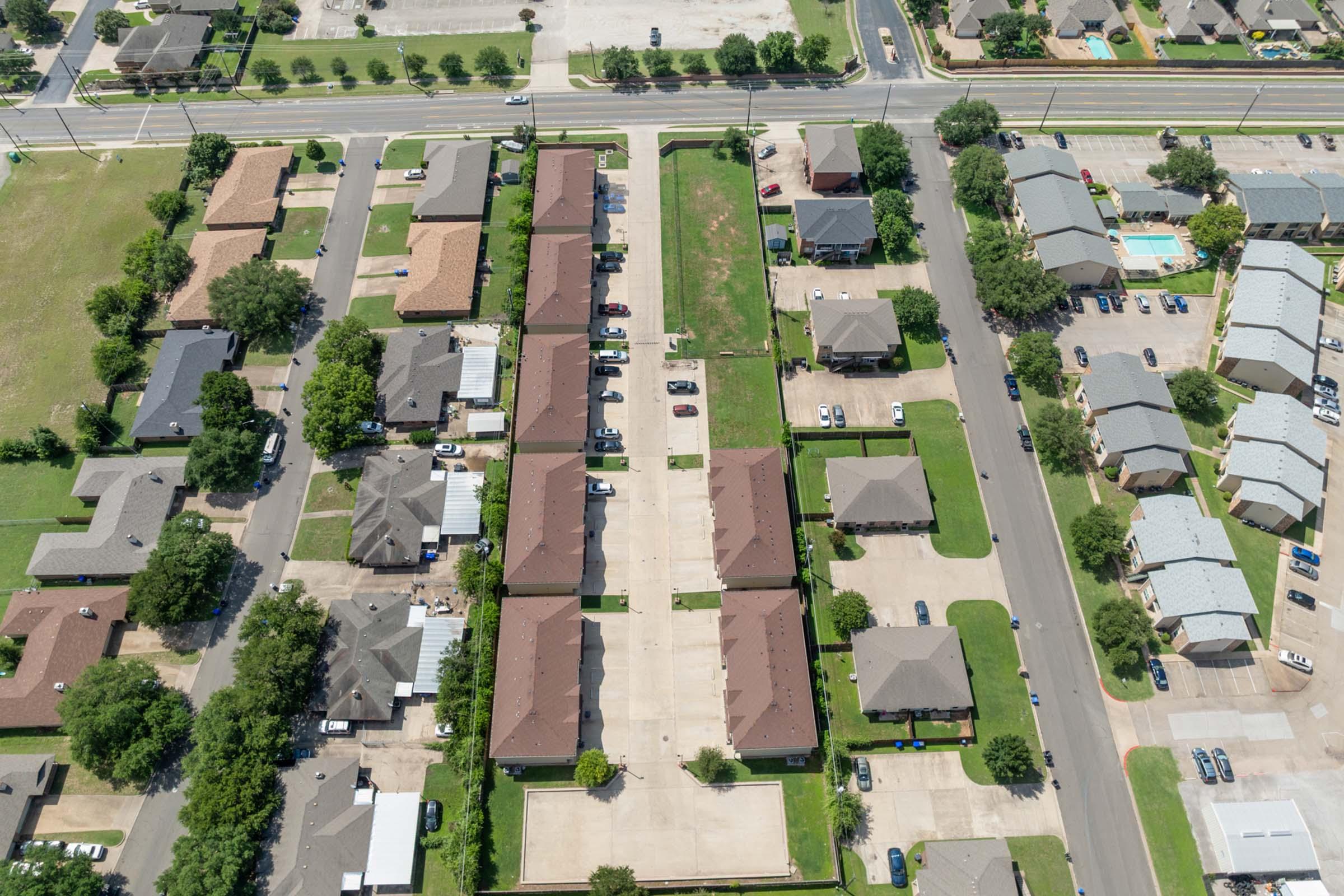 Aerial view of a neighborhood featuring rows of houses with brown roofs, surrounded by green lawns and parking spaces. Streets are lined with additional buildings, including an apartment complex and a pool area, set in a suburban landscape.