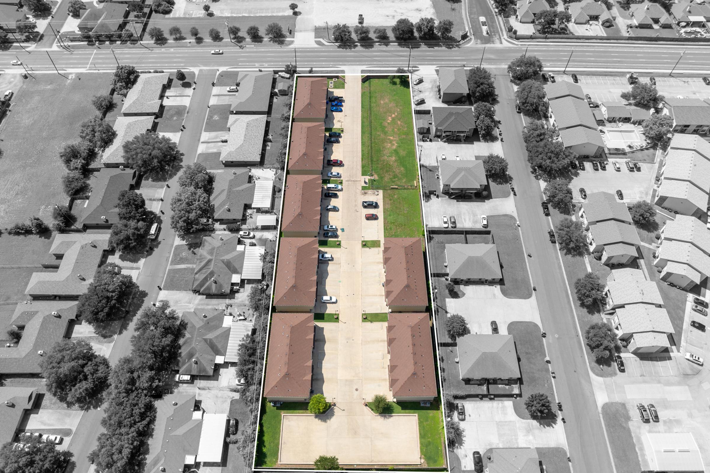 Aerial view of a residential area featuring multiple buildings with rooftops in a brownish hue. The parking lot is visible, with several cars parked. Surrounding the buildings are grassy areas and trees. The surrounding neighborhood includes more homes and roads. The image has a predominantly black-and-white appearance, highlighting the buildings.