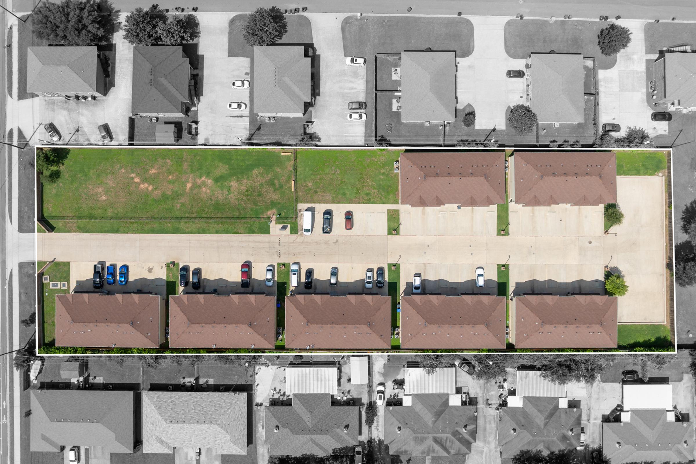 Aerial view of a residential area featuring multiple single-story buildings with brown roofs, arranged in rows. A grassy area is visible in the center, with several cars parked along the driveways. Surrounding homes and a street can be seen at the edges of the image.