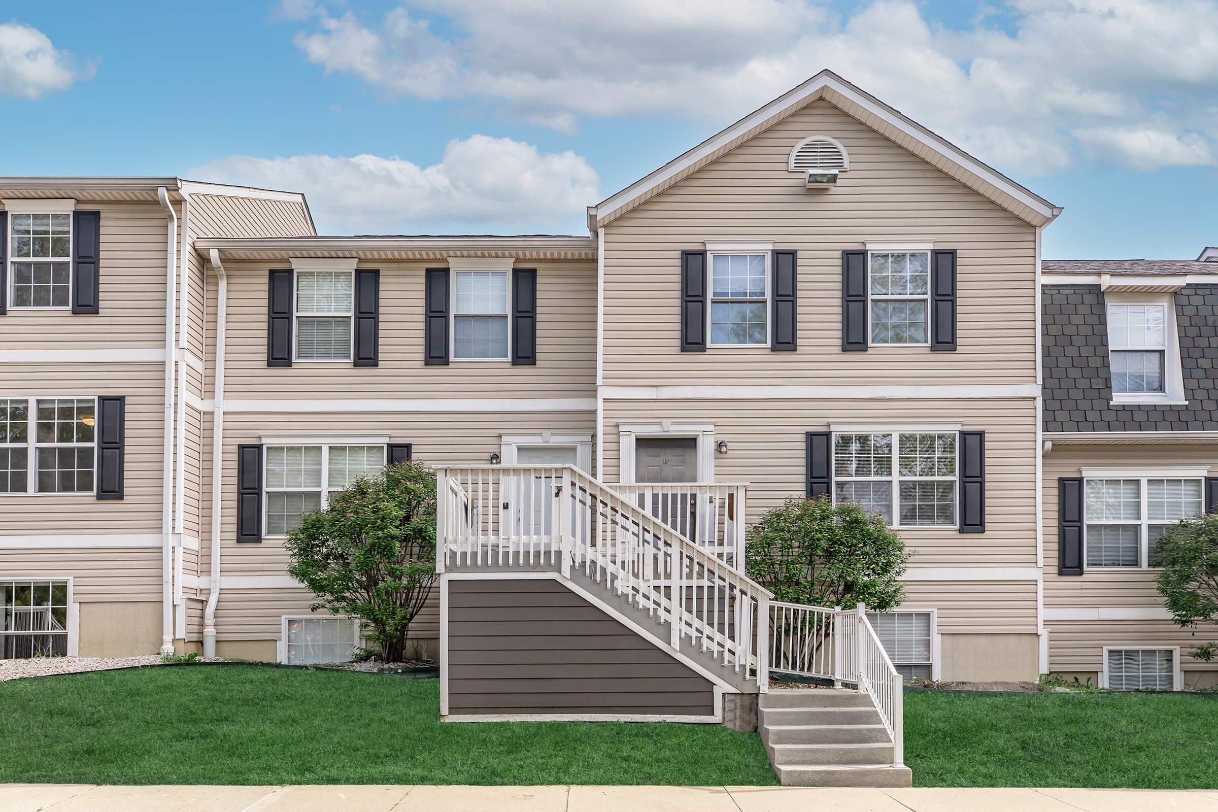 Two-story residential building with beige siding and black shutters. The building features multiple windows, a prominent entrance with stairs leading up, and landscaped green grass in front. The sky above is partly cloudy, creating a serene suburban atmosphere.
