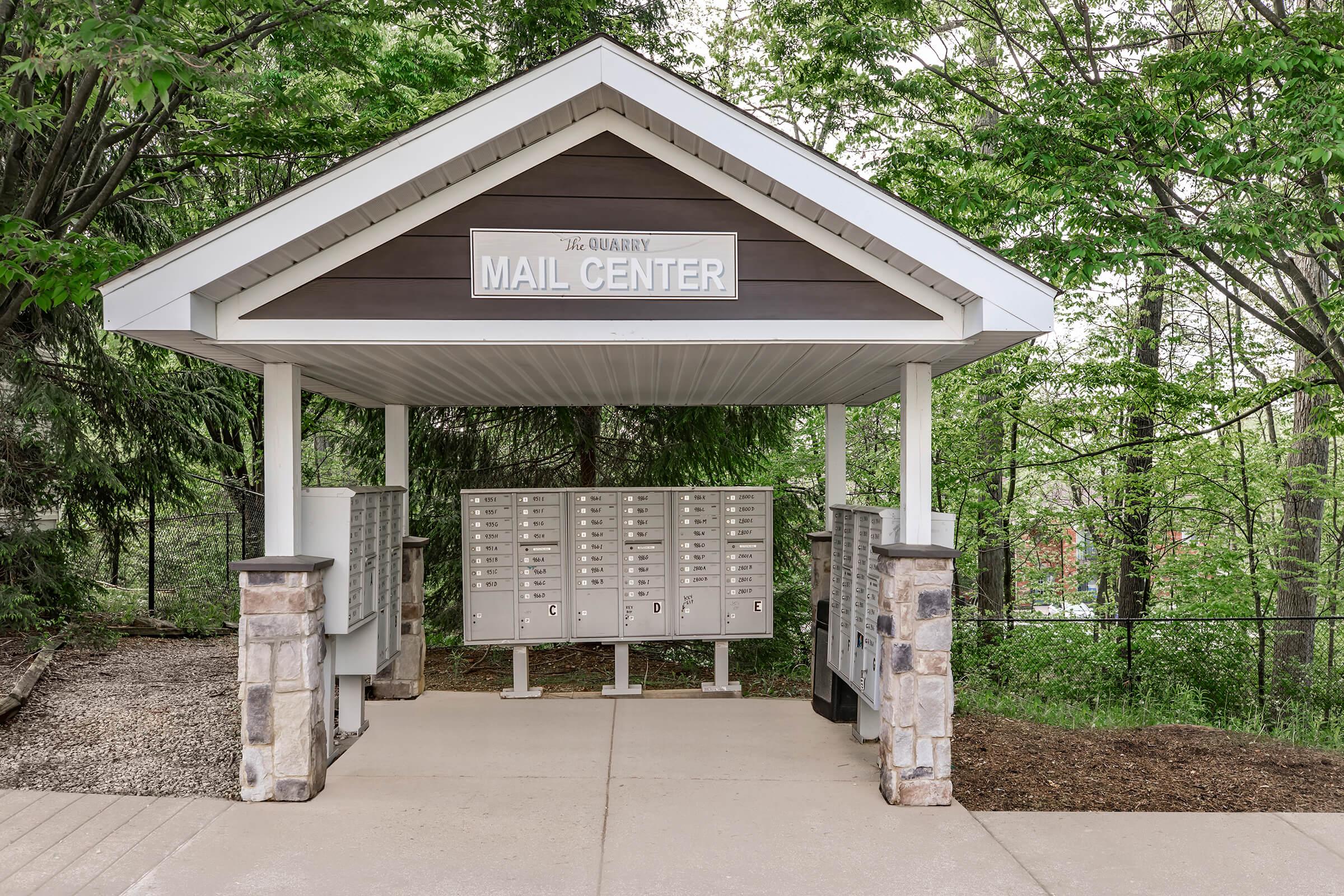 A mail center building with a peaked roof, featuring multiple mailboxes enclosed under a wooden structure. Surrounded by greenery and a paved walkway, the area is well-maintained and inviting. The sign above the entrance reads "The Quarry Mail Center."