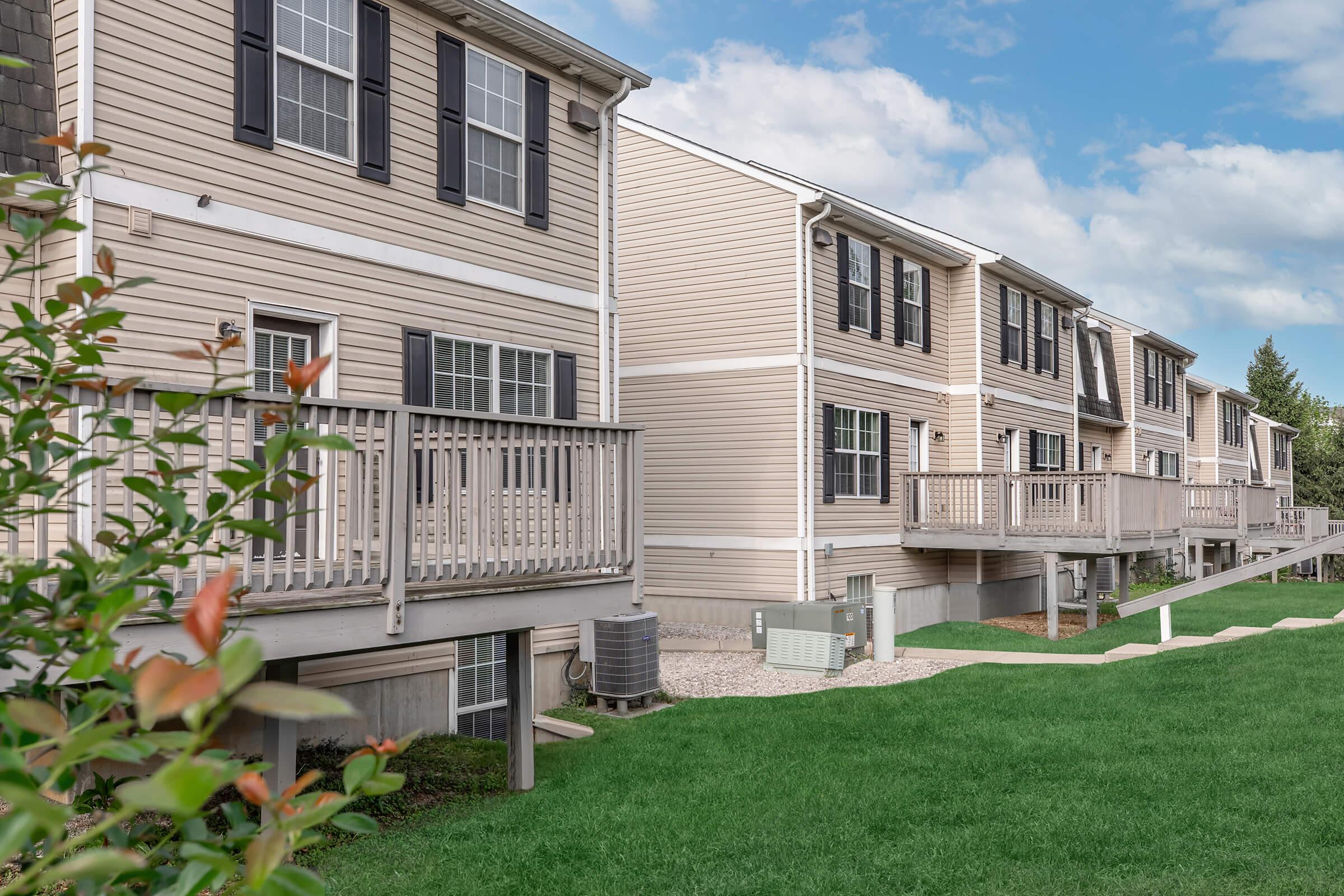 Row of residential buildings with beige siding, featuring balconies and large windows. The foreground includes a well-maintained green lawn and shrubbery, while air conditioning units are positioned near the base of the structures. The sky is blue with scattered clouds.