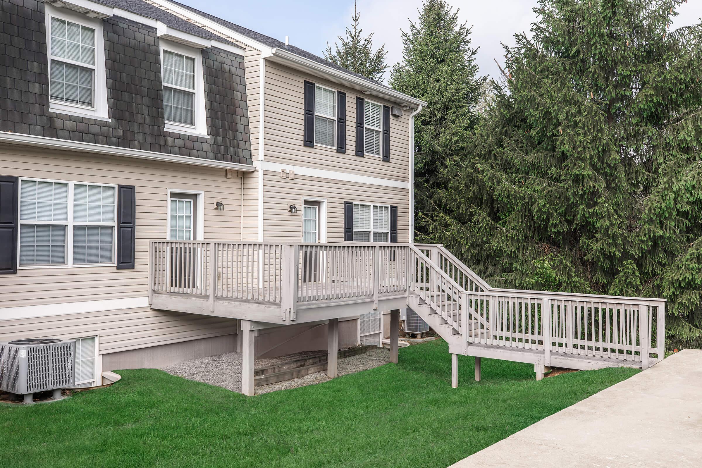 A two-story residential home with beige siding and dark shingles. There is a wooden deck with a railing on the side, leading to a set of stairs. The yard is grassy, with a small air conditioning unit visible on the left side of the house. Tall evergreen trees are in the background.