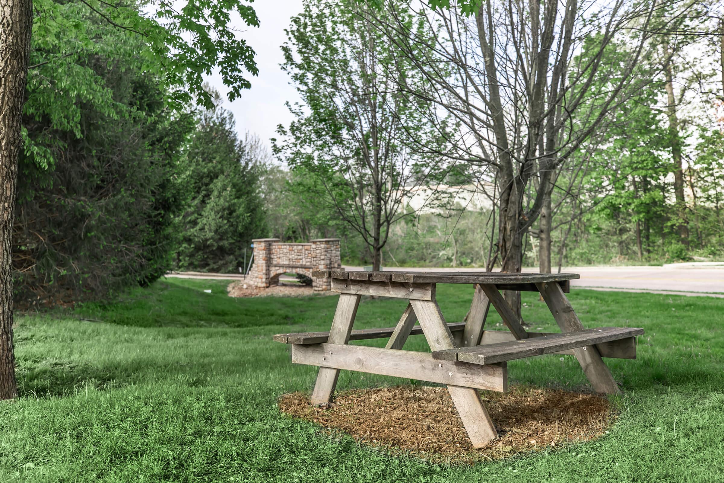 A wooden picnic table sits on grassy terrain surrounded by trees. In the background, a brick fireplace is visible, adding to the natural outdoor setting. The scene is tranquil, suggesting a perfect spot for outdoor gatherings or relaxation.