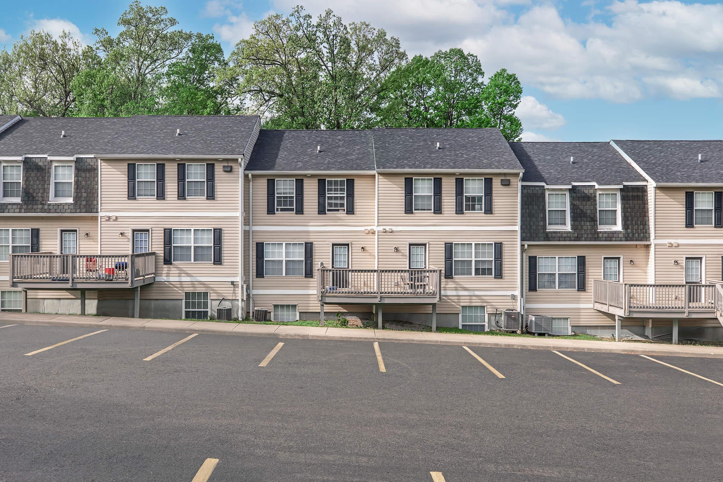 Row of residential apartments featuring multiple units, each with balconies and a tree-lined background. The pavement in front has designated parking spaces, and the sky is partly cloudy, indicating a pleasant day.