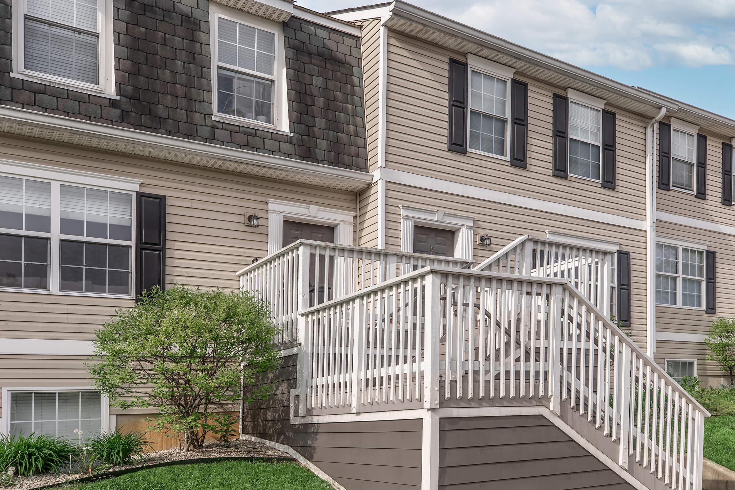 Two townhouses with a front staircase leading to the main entrances. The buildings feature alternating siding colors, with windows dressed in black shutters. Lush greenery surrounds the base, and a clear sky is visible above, adding a bright and inviting atmosphere.