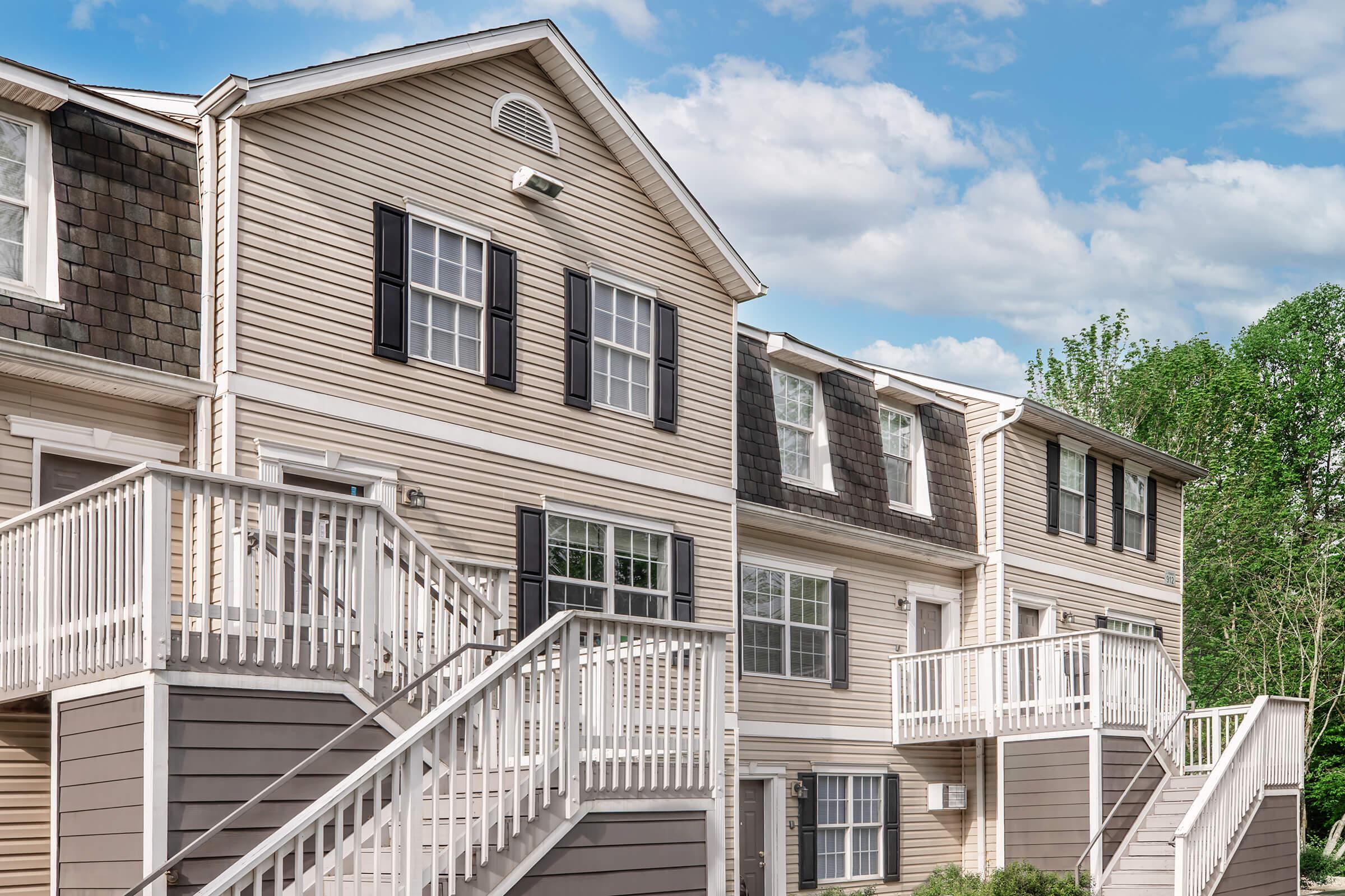 A row of residential apartment units featuring beige siding and black shutters. Each unit has a staircase leading to a front door, with green trees in the background against a partly cloudy sky. The architecture reflects a modern suburban style.