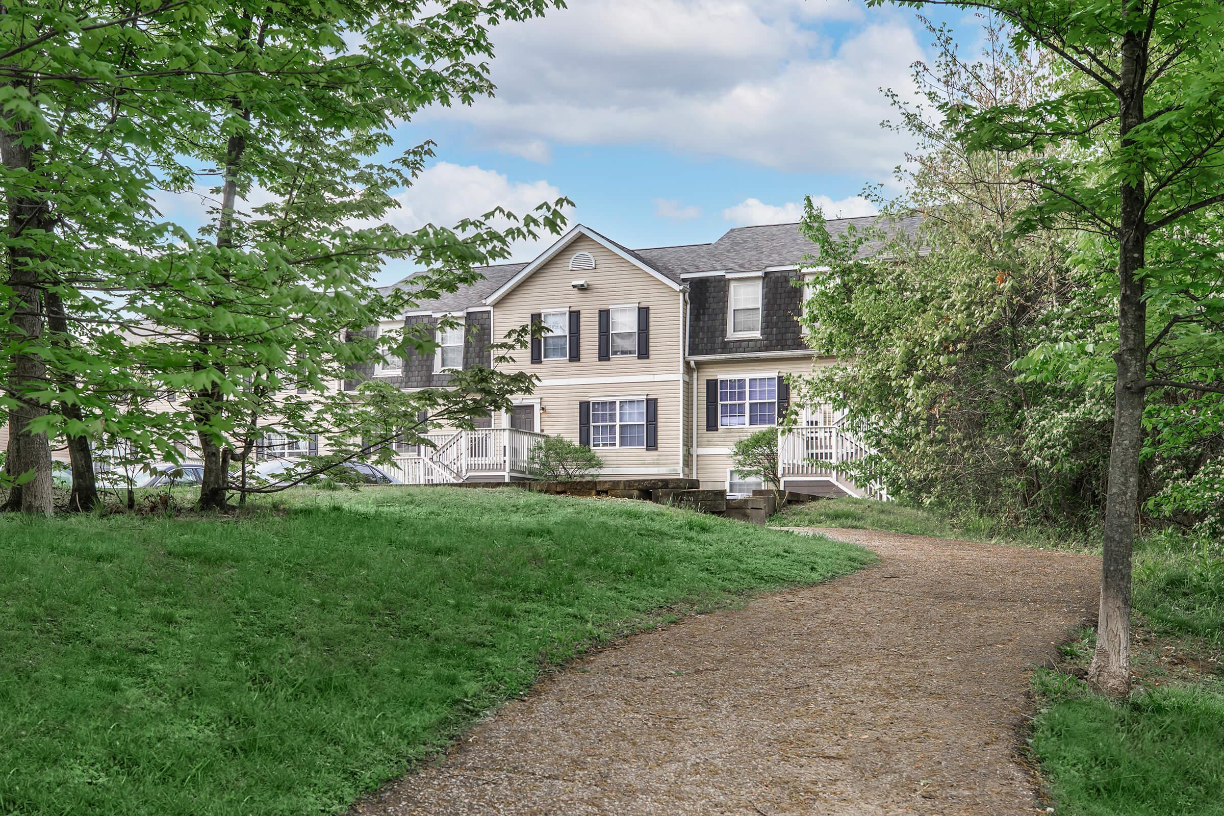 A serene pathway leads to a charming two-story house surrounded by lush green grass and trees. The house features a light-colored exterior with multiple windows and a welcoming porch. Soft clouds dot the blue sky, enhancing the peaceful outdoor atmosphere.